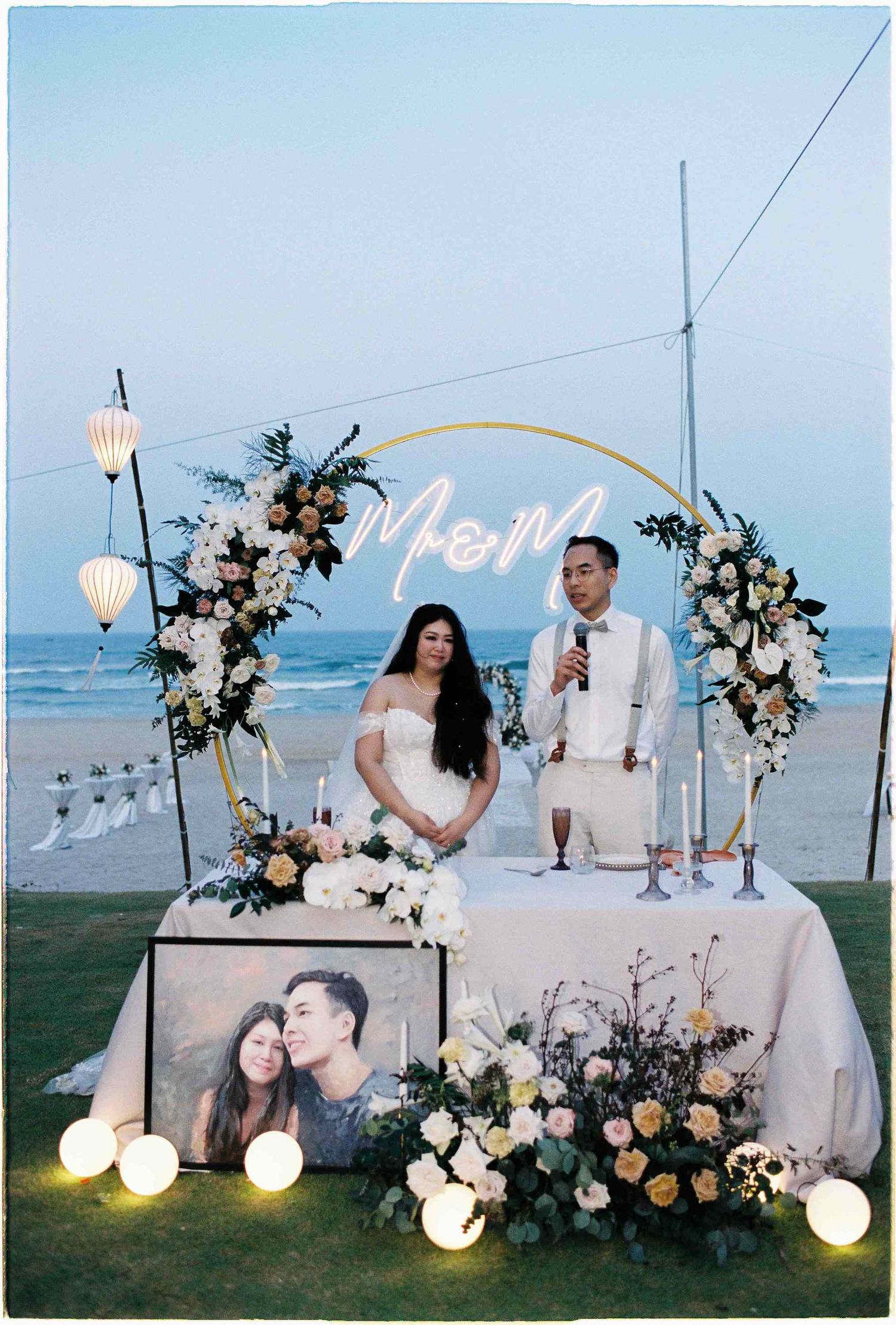 Couple at beach wedding reception table with floral arch, neon M&M sign, candles, and framed photo at sunset.