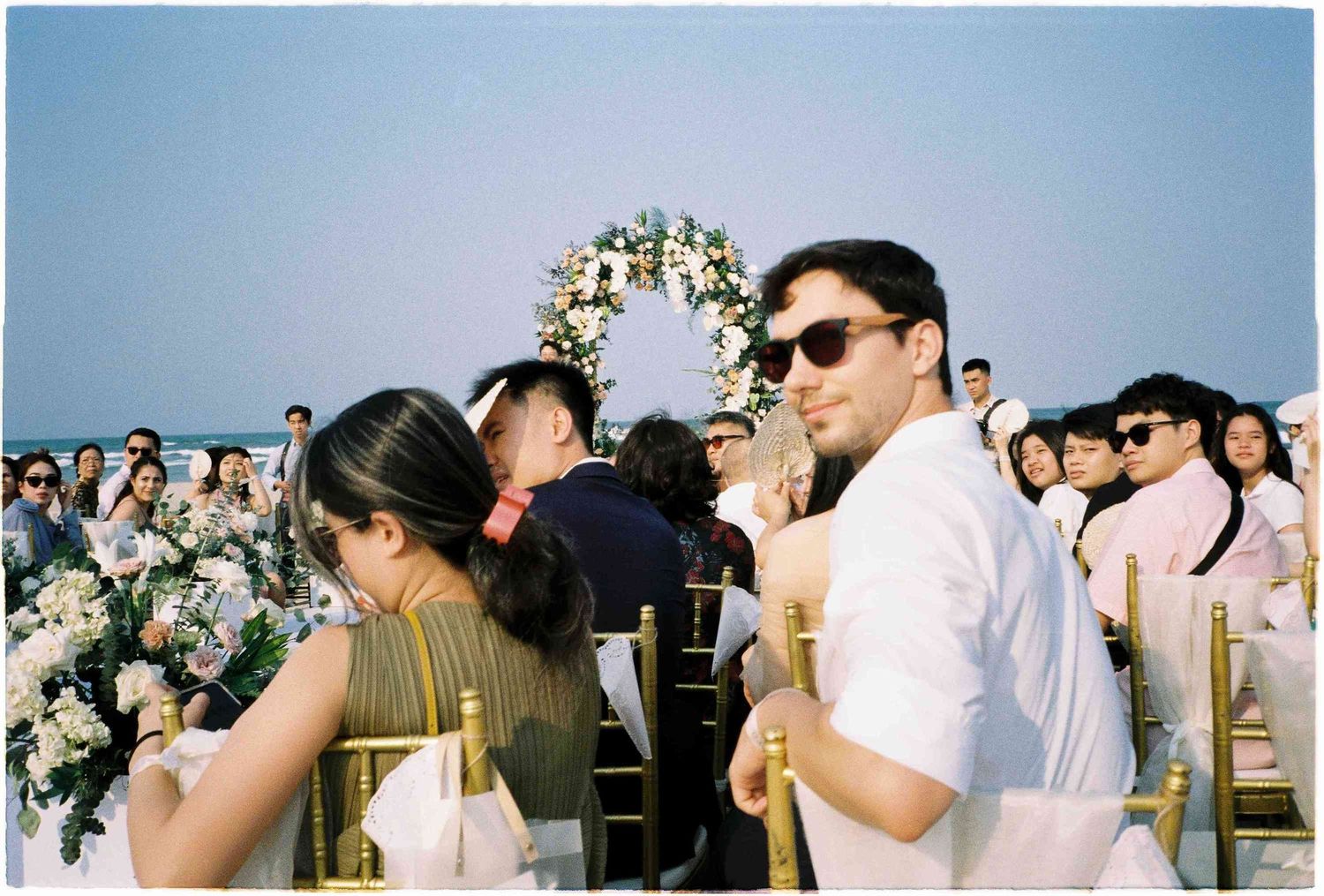 Outdoor beach wedding ceremony with guests seated on gold chairs, floral arch, and a man in white suit wearing sunglasses.