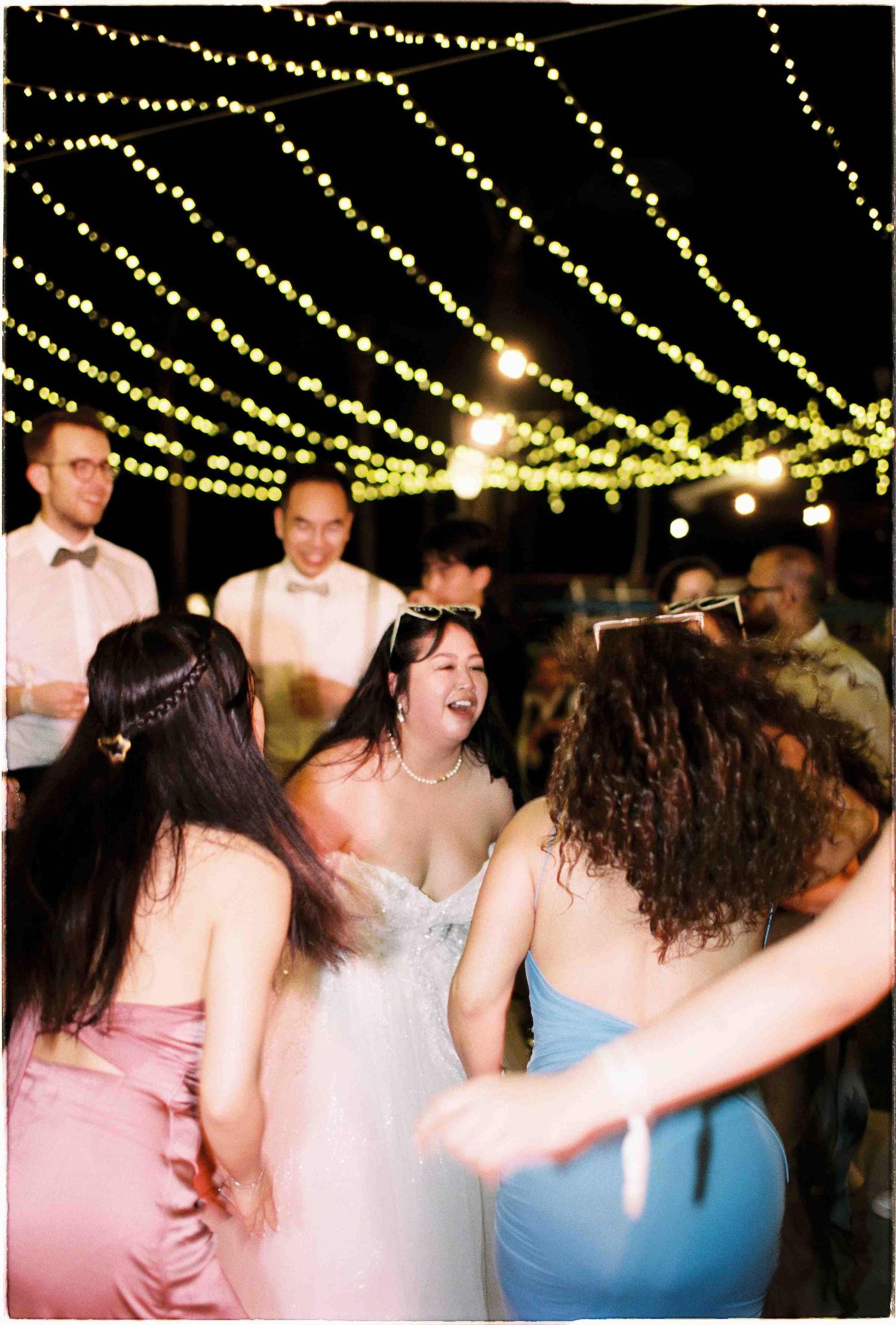 Bride laughing joyfully with bridesmaids dancing under string lights at outdoor wedding reception at night.