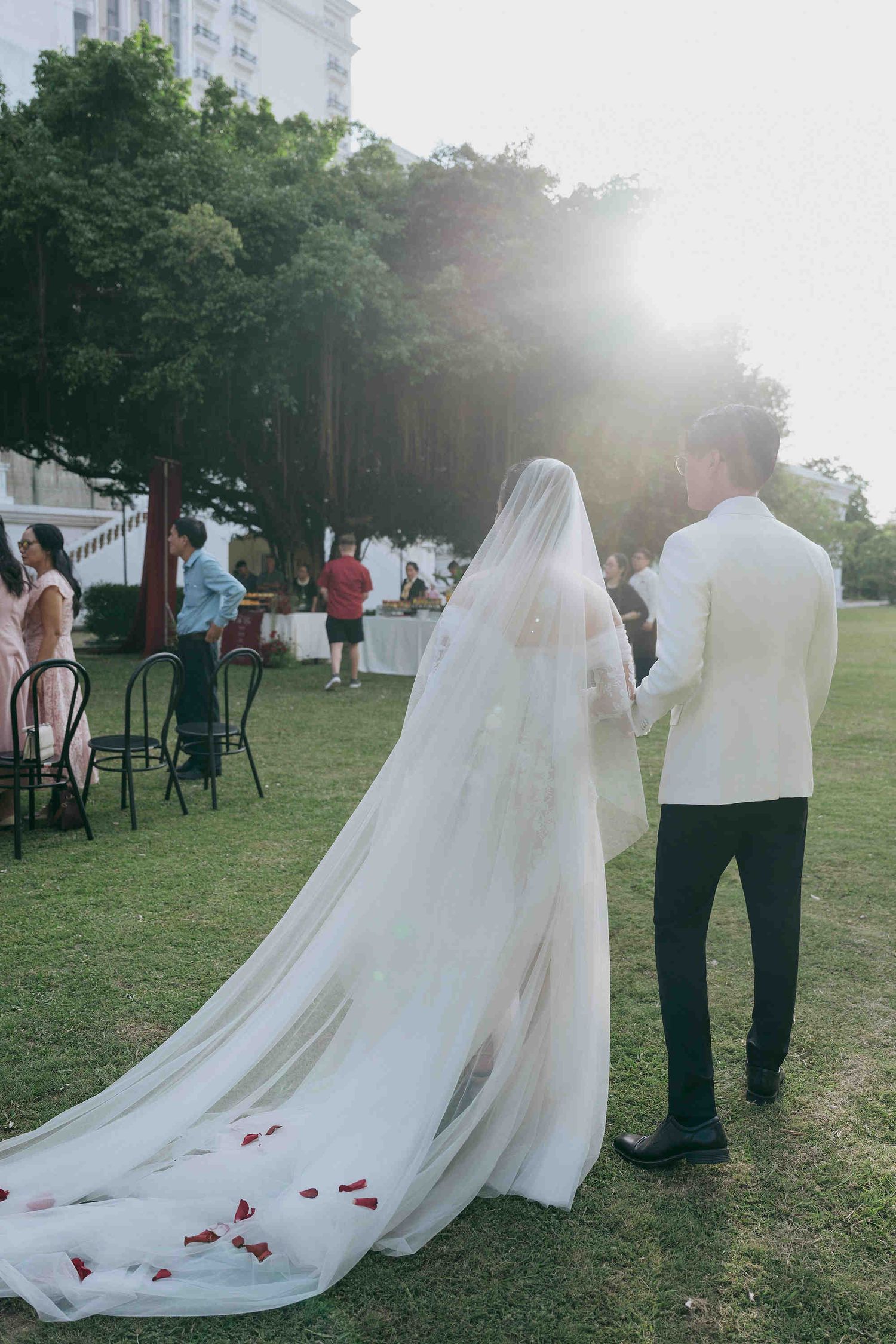 Bride in white gown with floral veil and groom in white suit walking on green lawn at outdoor wedding.