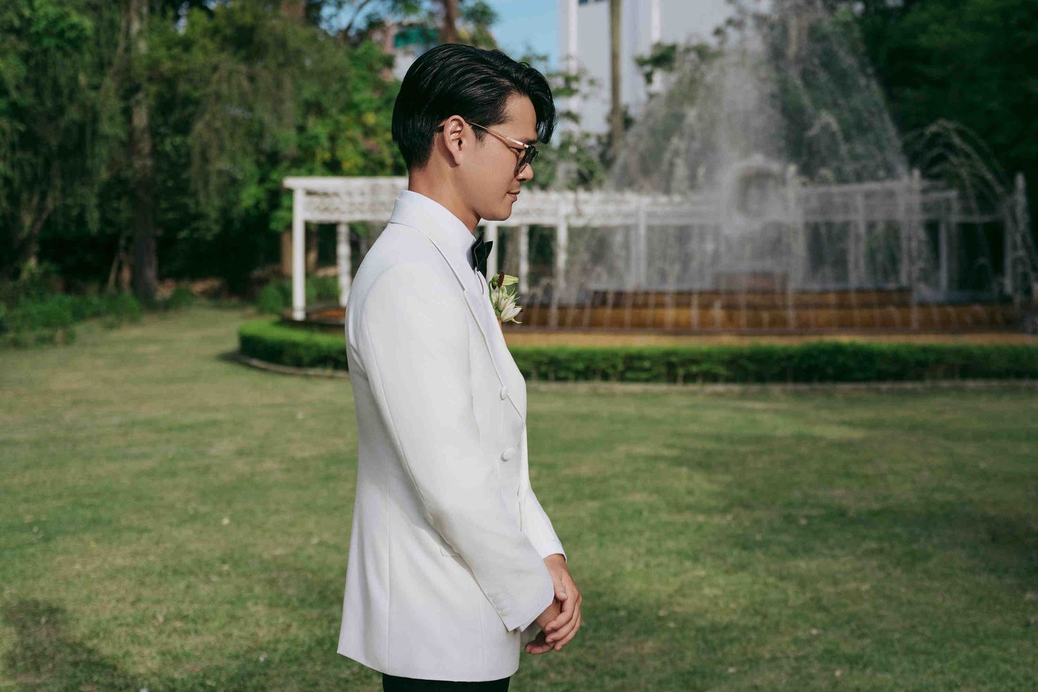 A groom in a white tuxedo jacket with boutonniere stands in a garden with a fountain backdrop.