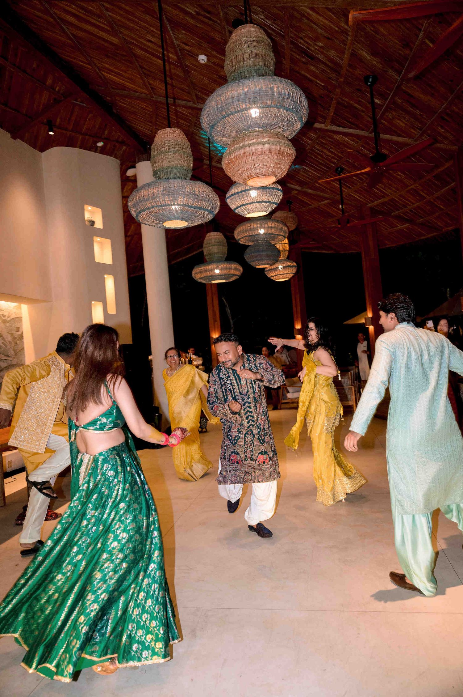 Guests in vibrant Indian attire dance under woven pendant lights at a festive celebration.