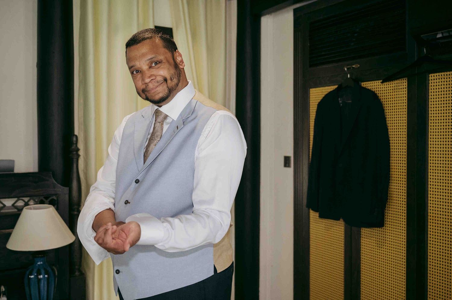 Man in light blue vest and white shirt smiling while adjusting cufflinks indoors.