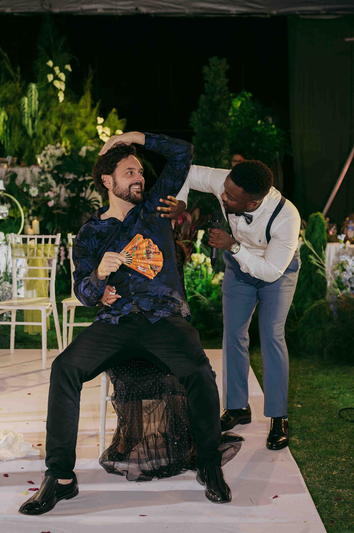 Two men celebrate at an outdoor wedding reception, one seated holding a fan while the other dances behind him.