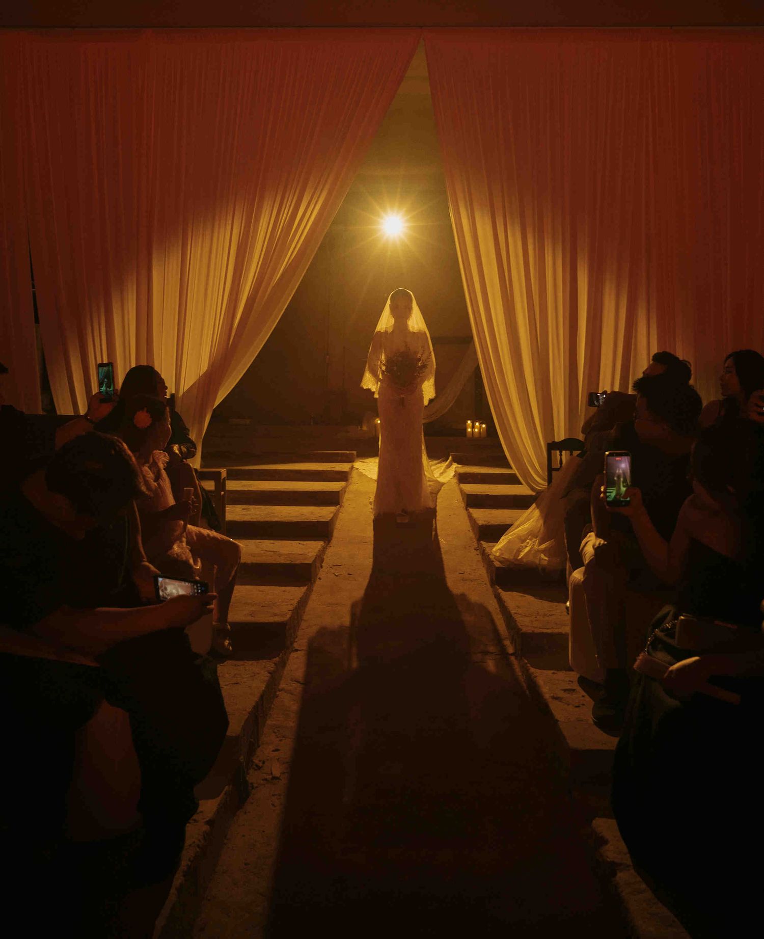 Bride in white gown and veil walks down a dramatic candlelit aisle flanked by curtains and seated guests.