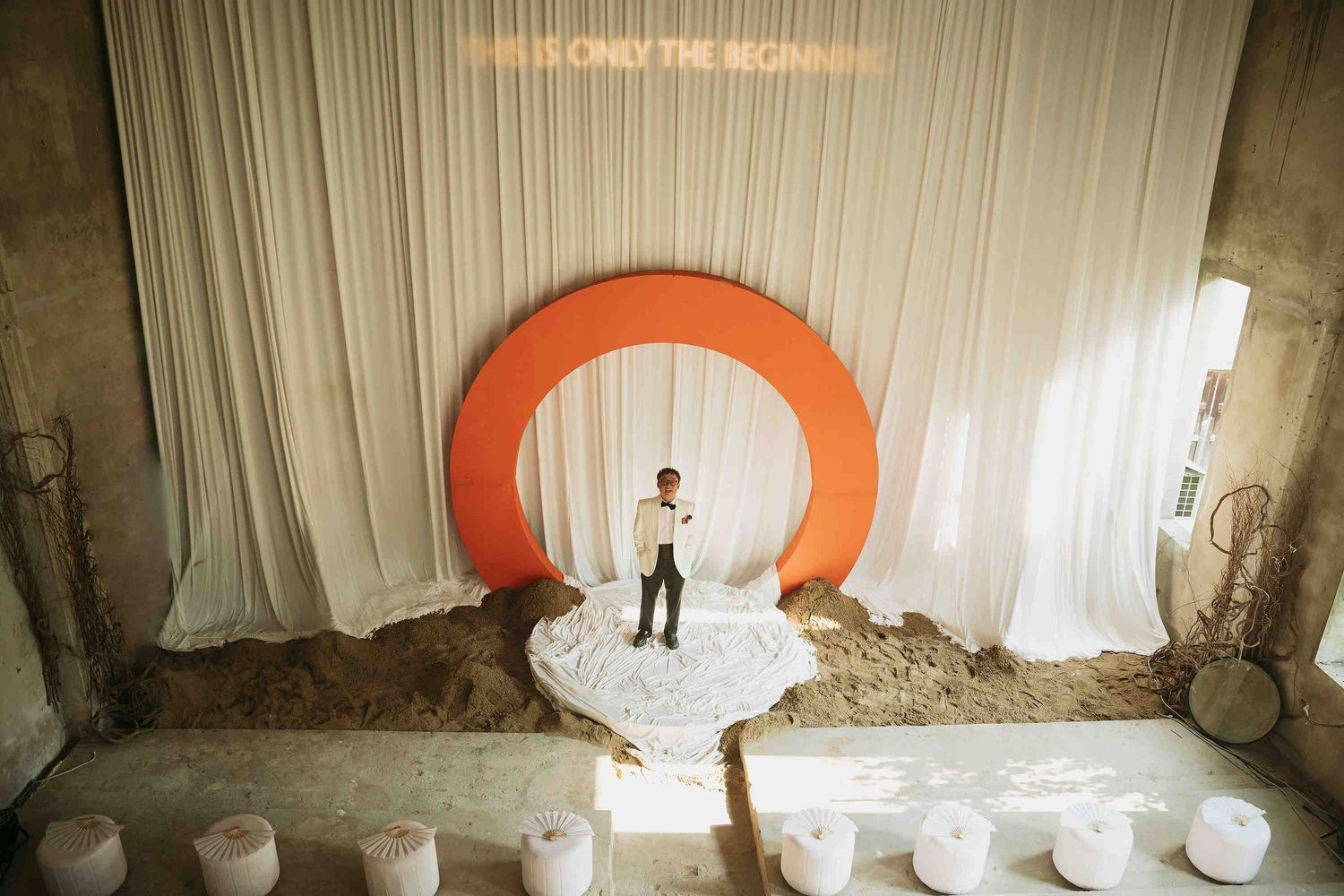 Person standing before a large orange circular arch installation with white draping and neon sign backdrop.