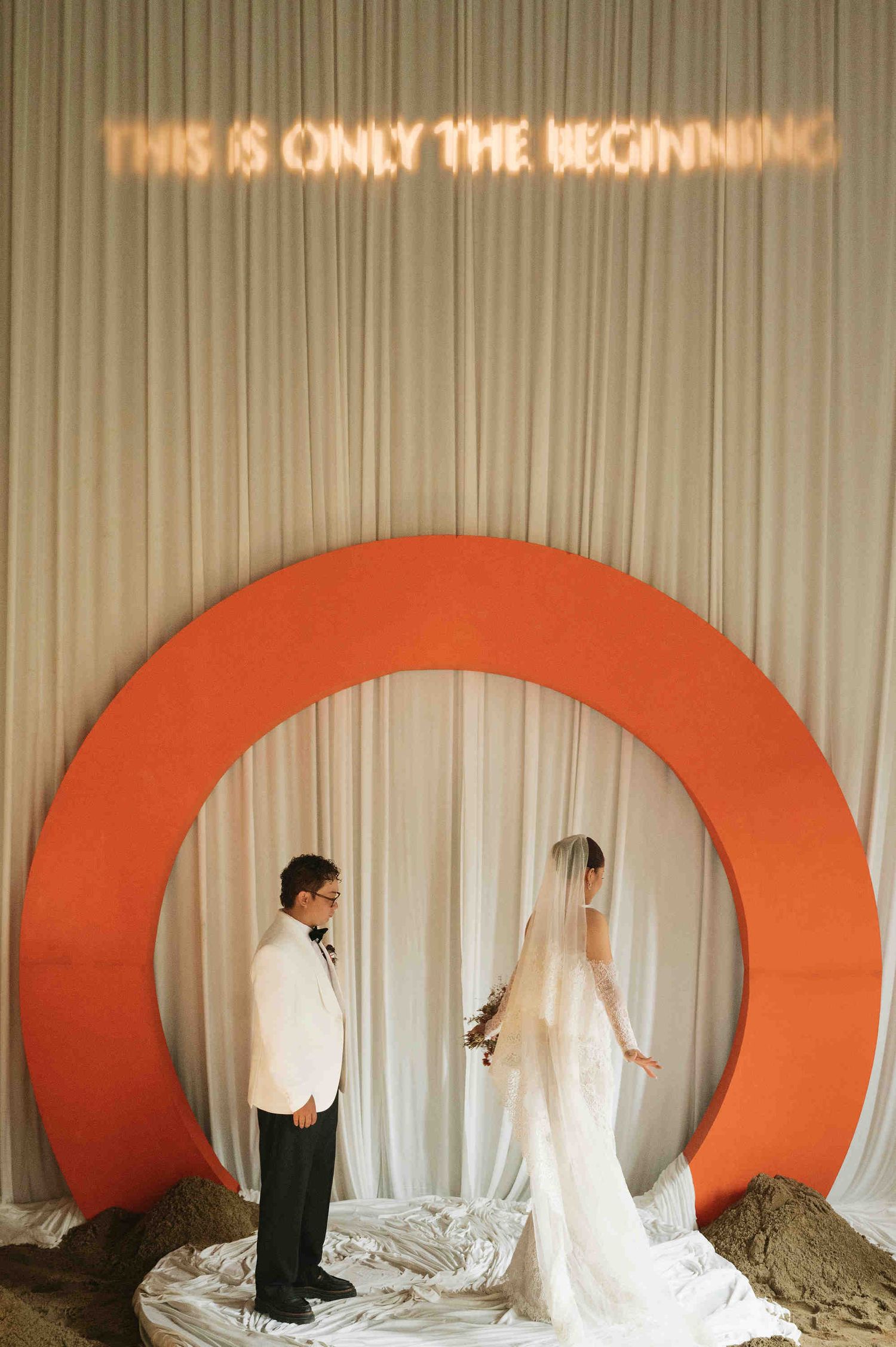 Bride and groom stand before an orange circular arch with neon sign reading This Is Only The Beginning.