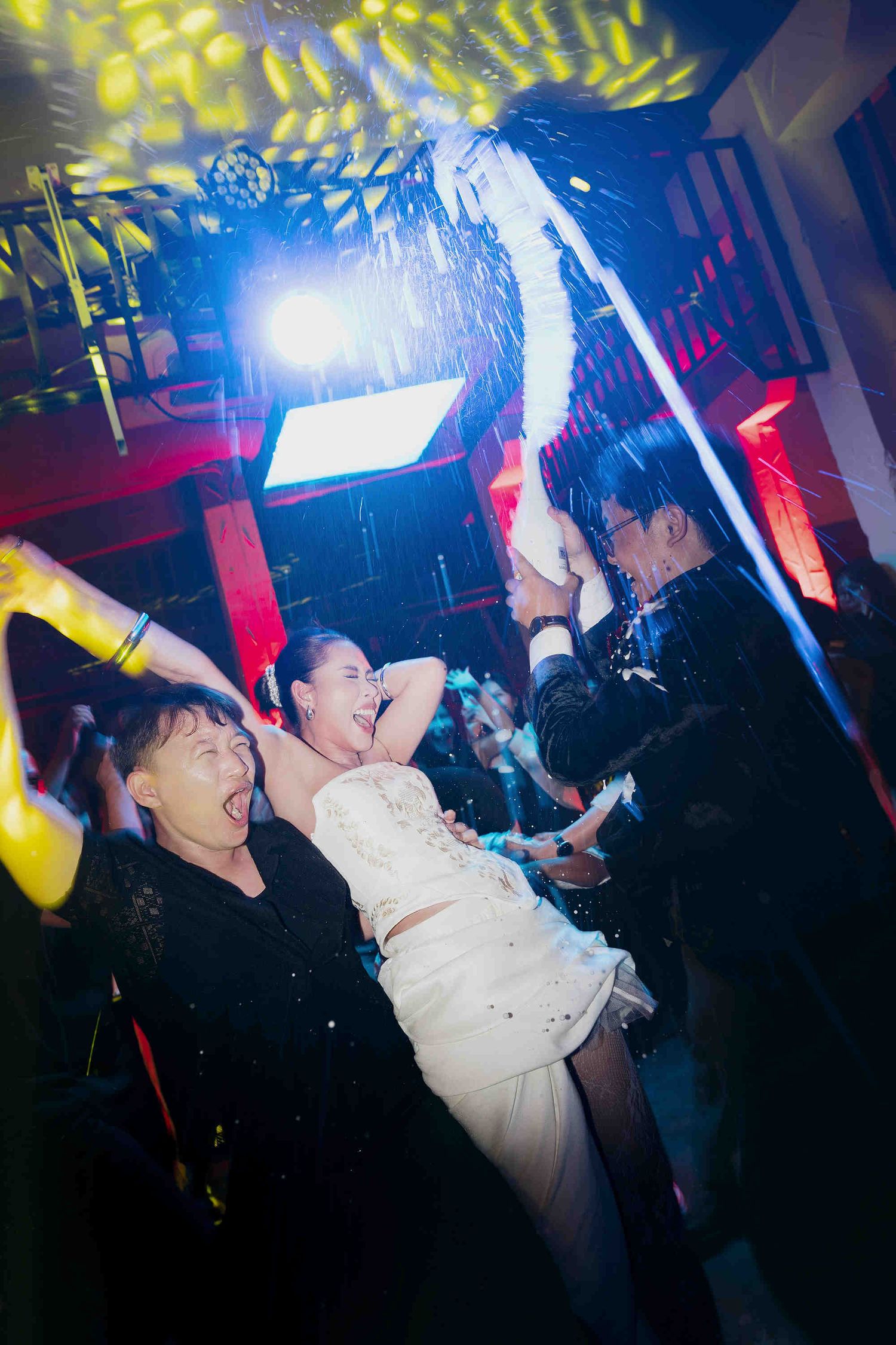 Joyful wedding party celebration on dance floor with confetti, bride in white dress surrounded by guests under colorful lights.