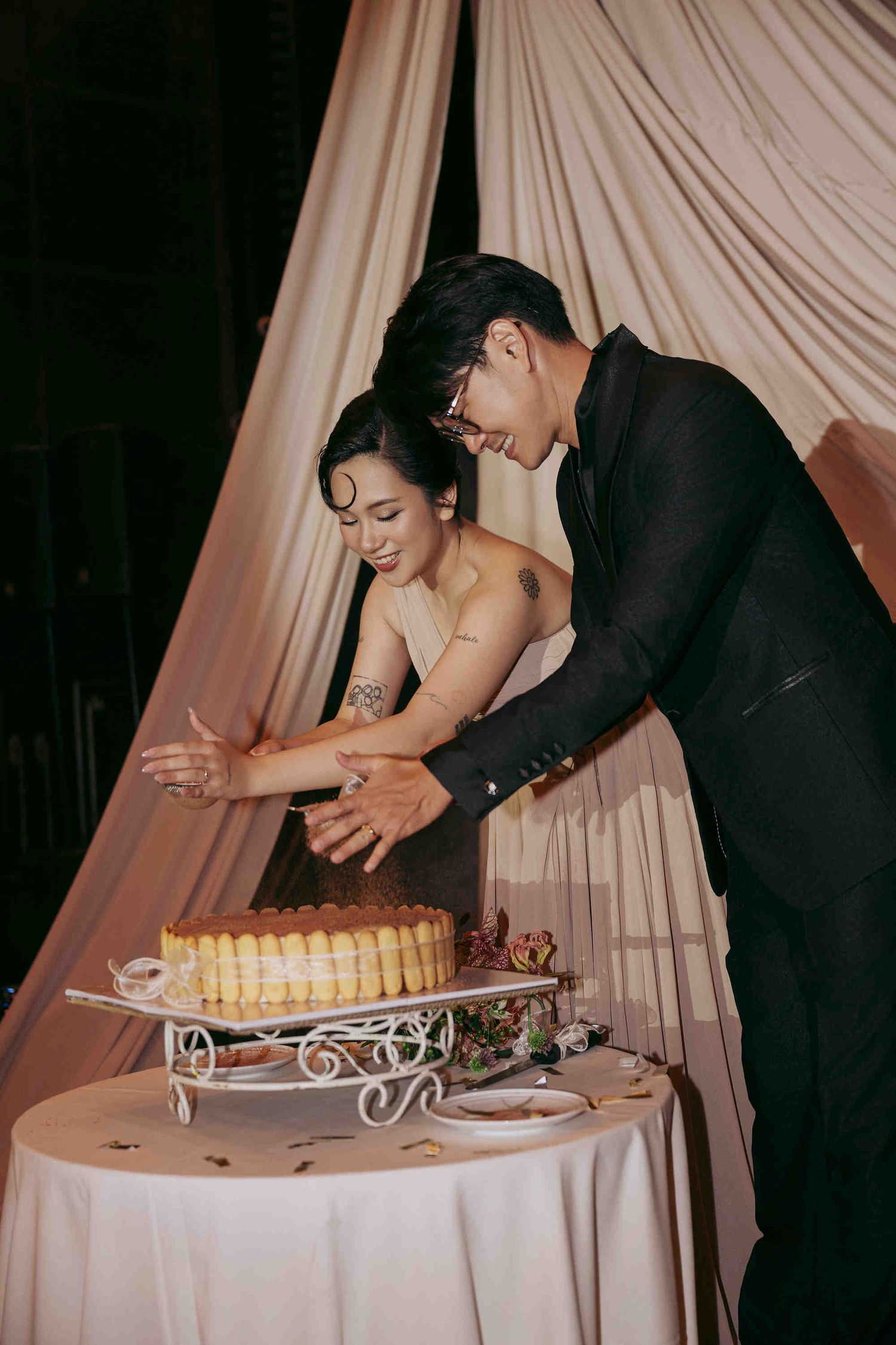 Couple cutting wedding cake together at elegant reception, woman in strapless gown and man in black suit, soft draped backdrop.