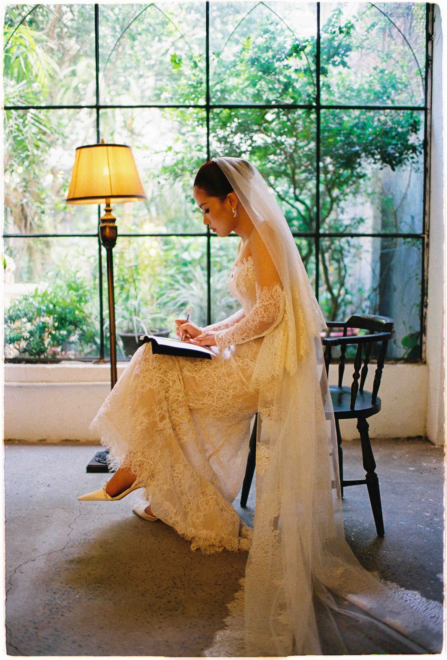 Bride in lace wedding gown and veil writing in a journal by a floor lamp near large greenhouse windows.