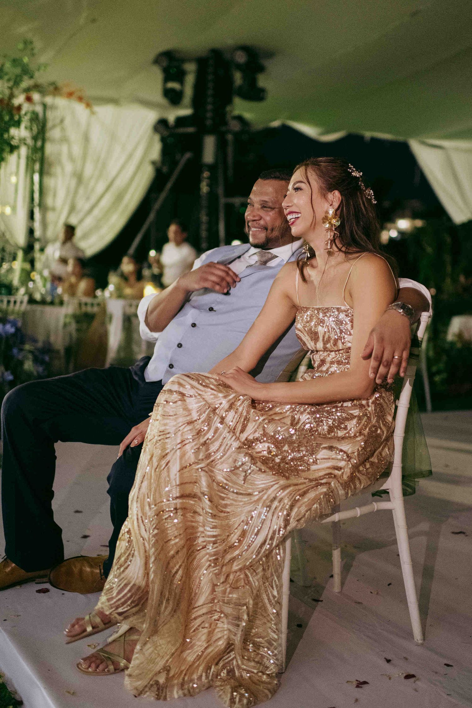 Couple in elegant attire smiling and seated together at a wedding reception under a decorated tent.
