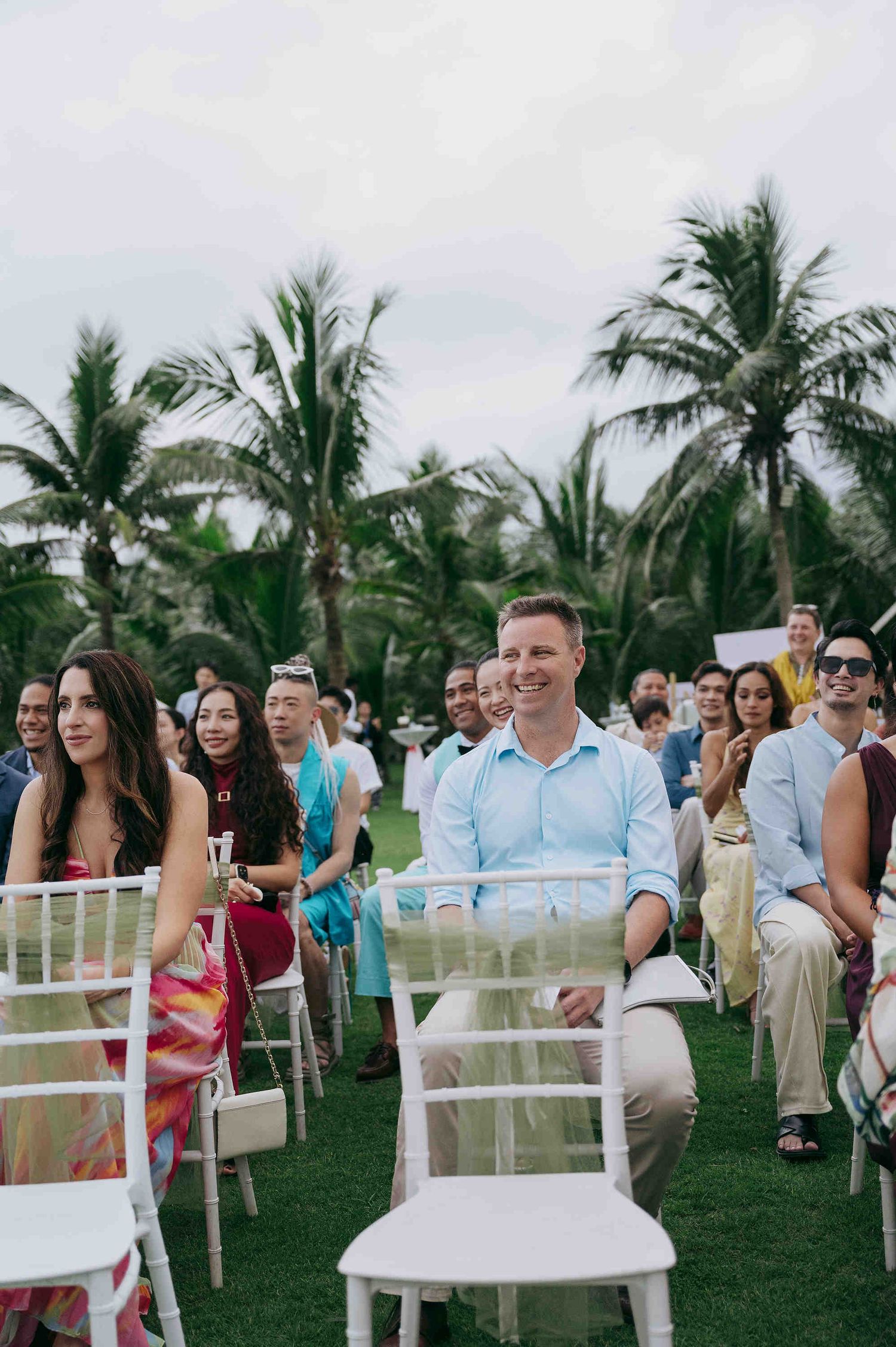 Guests seated outdoors at a tropical wedding ceremony with palm trees in the background.