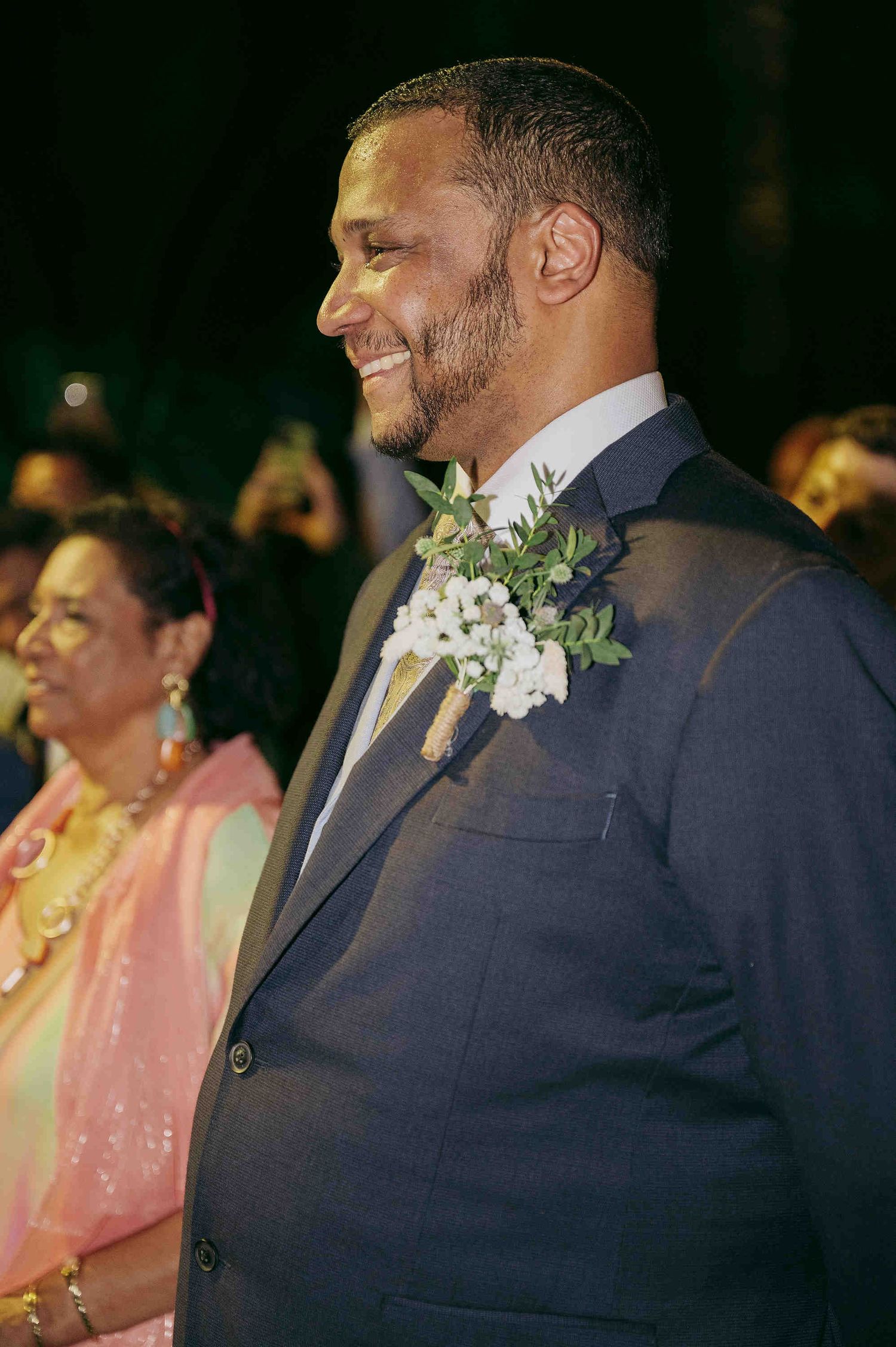 Smiling groom in navy suit with white floral boutonniere at elegant wedding celebration.