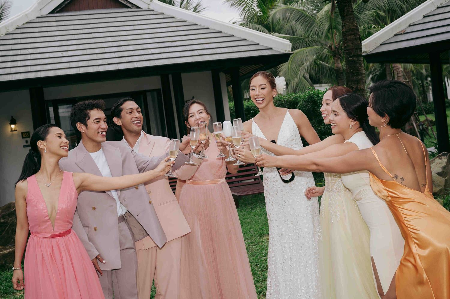 A joyful wedding party toasts with champagne glasses outdoors, featuring a bride in white and guests in pastel gowns.