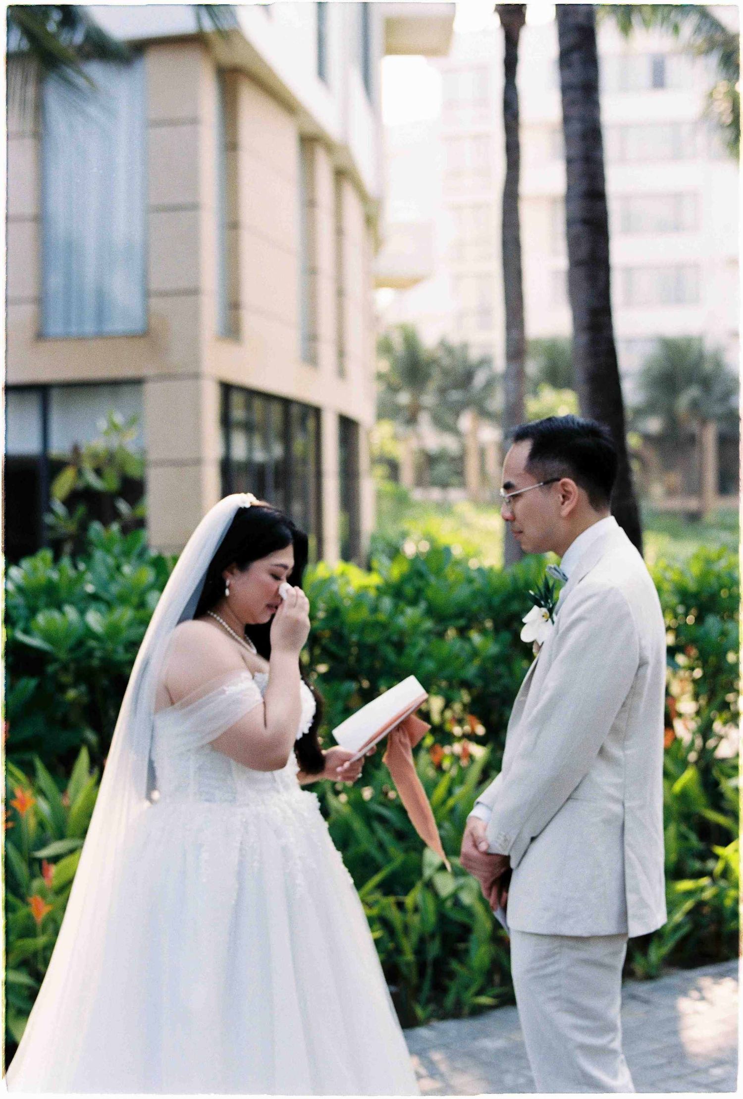 Emotional bride reads vows while groom listens at outdoor wedding ceremony surrounded by tropical greenery.