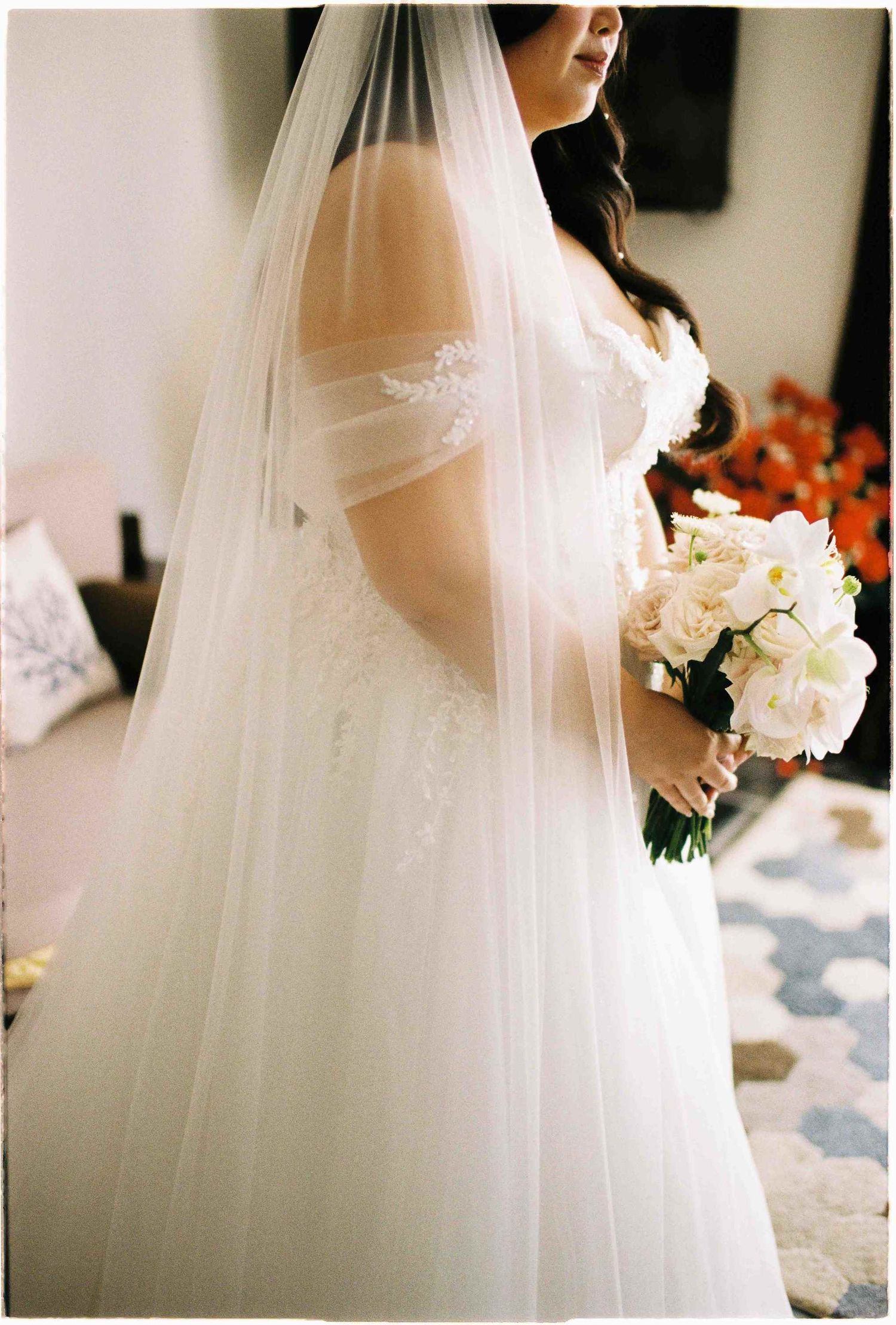 Bride in off-shoulder lace wedding gown with flowing veil holding white and blush rose bouquet.