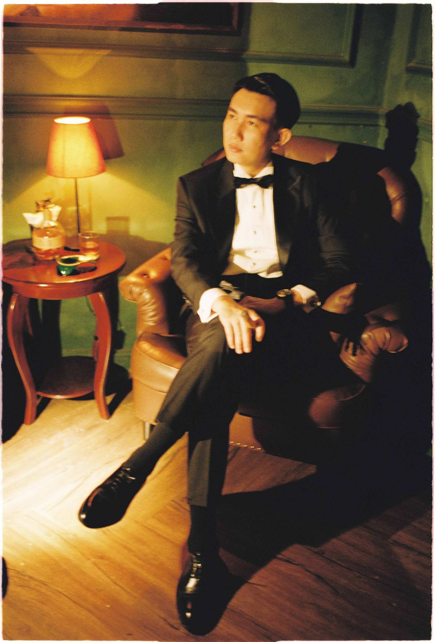 Vietnam pre-wedding: man in black tuxedo seated in leather armchair with drink and table lamp, by AnhVuWedding.
