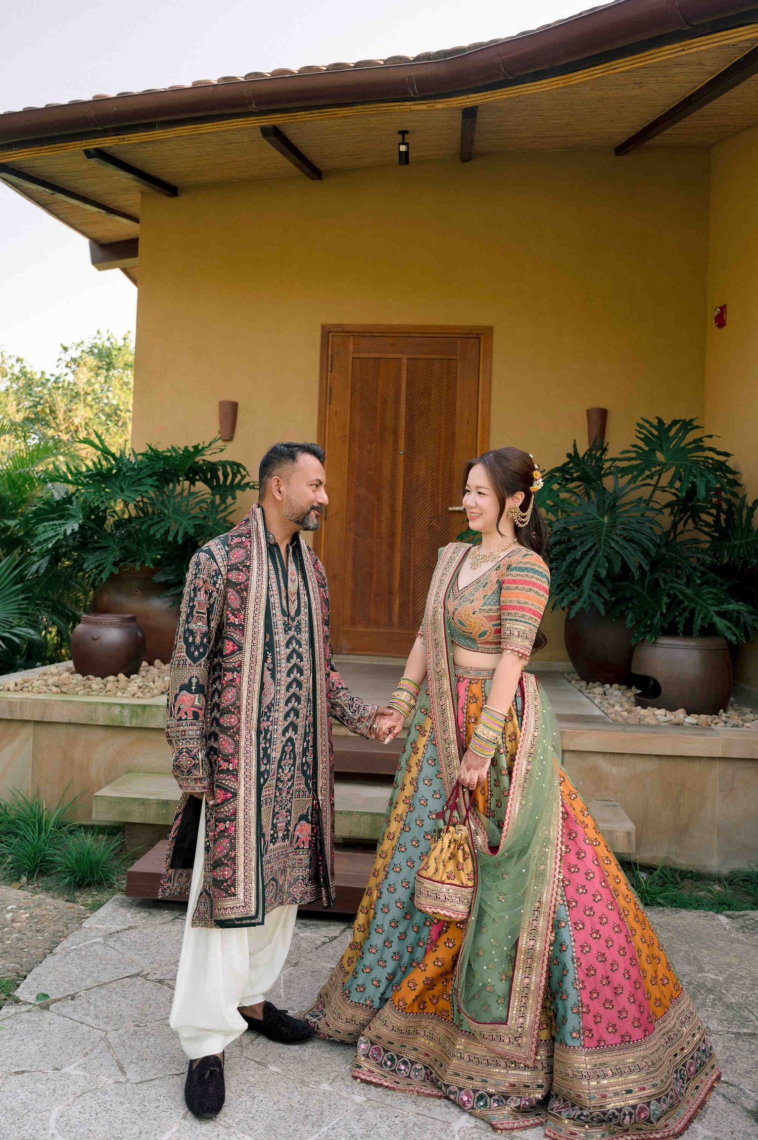 Couple in traditional Indian wedding attire holding hands outside a yellow villa with lush tropical greenery.