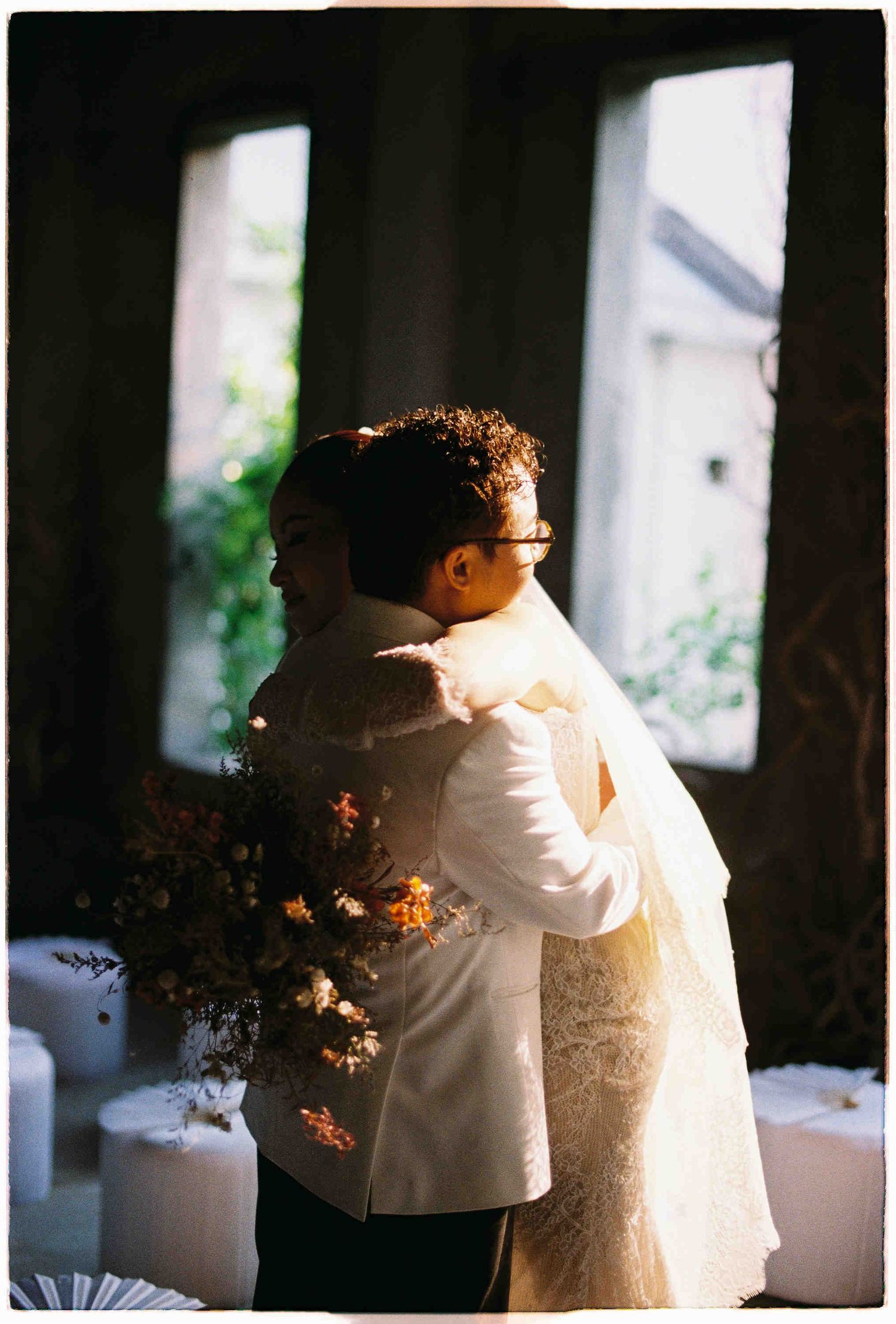 Bride and groom share a tender embrace in soft natural light, holding a floral bouquet at an elegant wedding venue.
