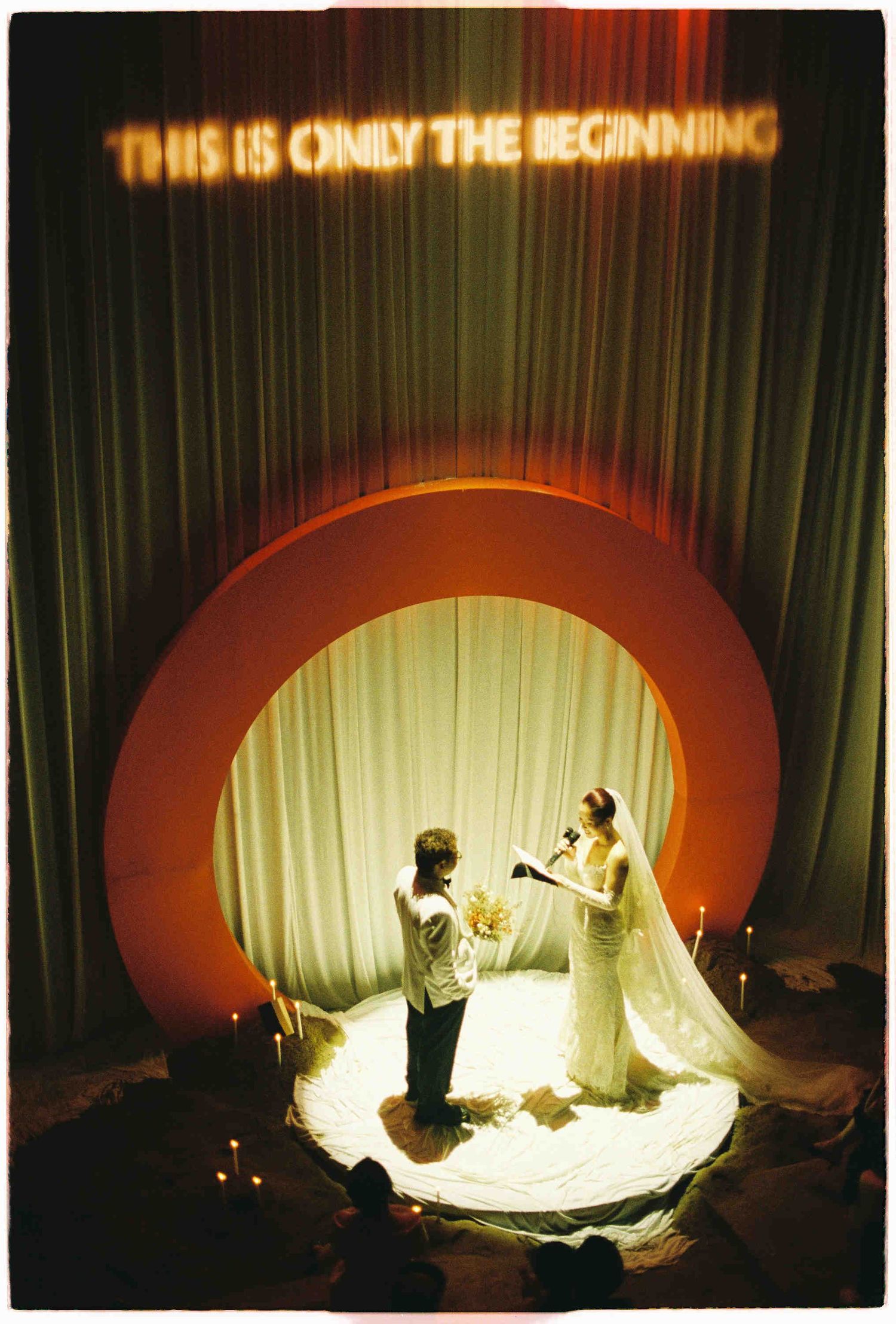 Couple exchanges vows under a circular arch with glowing text THIS IS ONLY THE BEGINNING at a candlelit wedding ceremony.