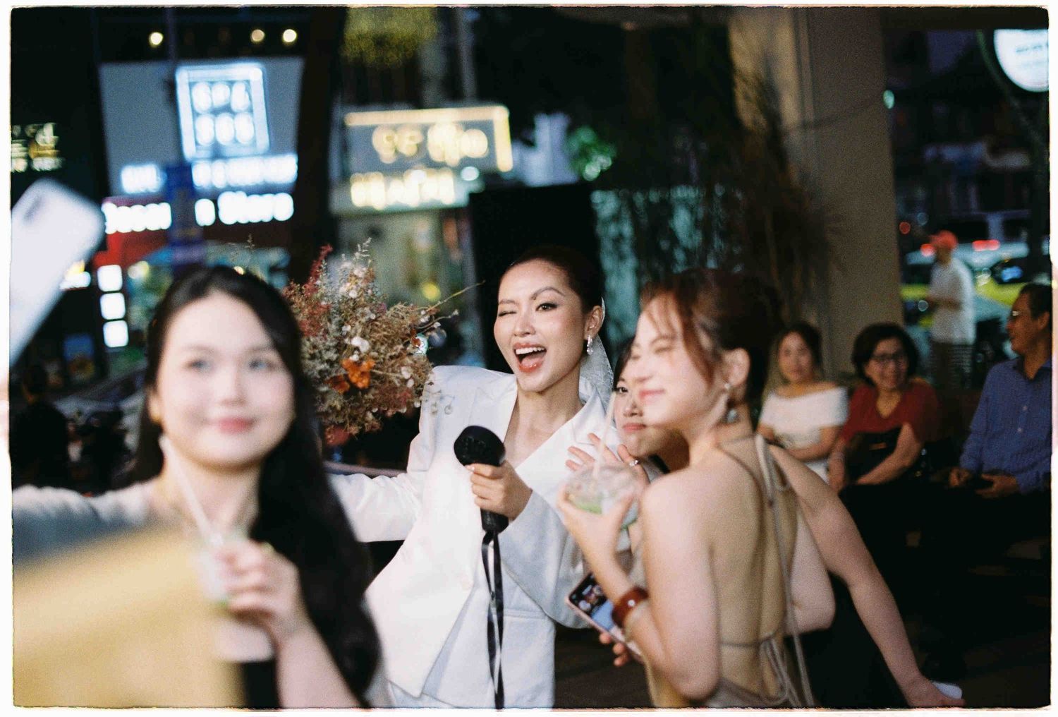 Women singing karaoke outdoors at a lively night event in an urban setting with bright city lights.