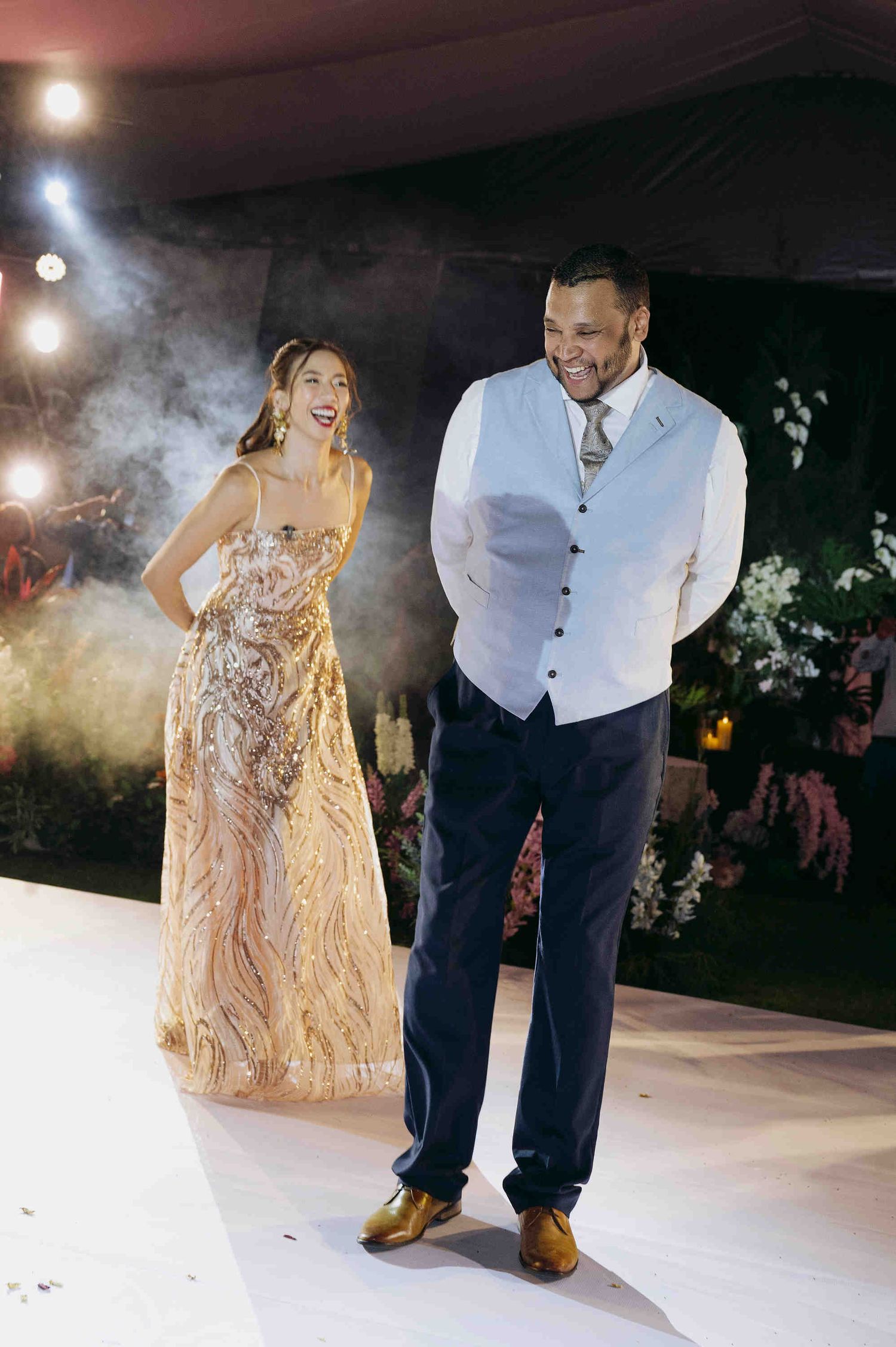 Couple in elegant formal attire walking at a wedding reception, woman in gold gown, man in vest and dress pants.