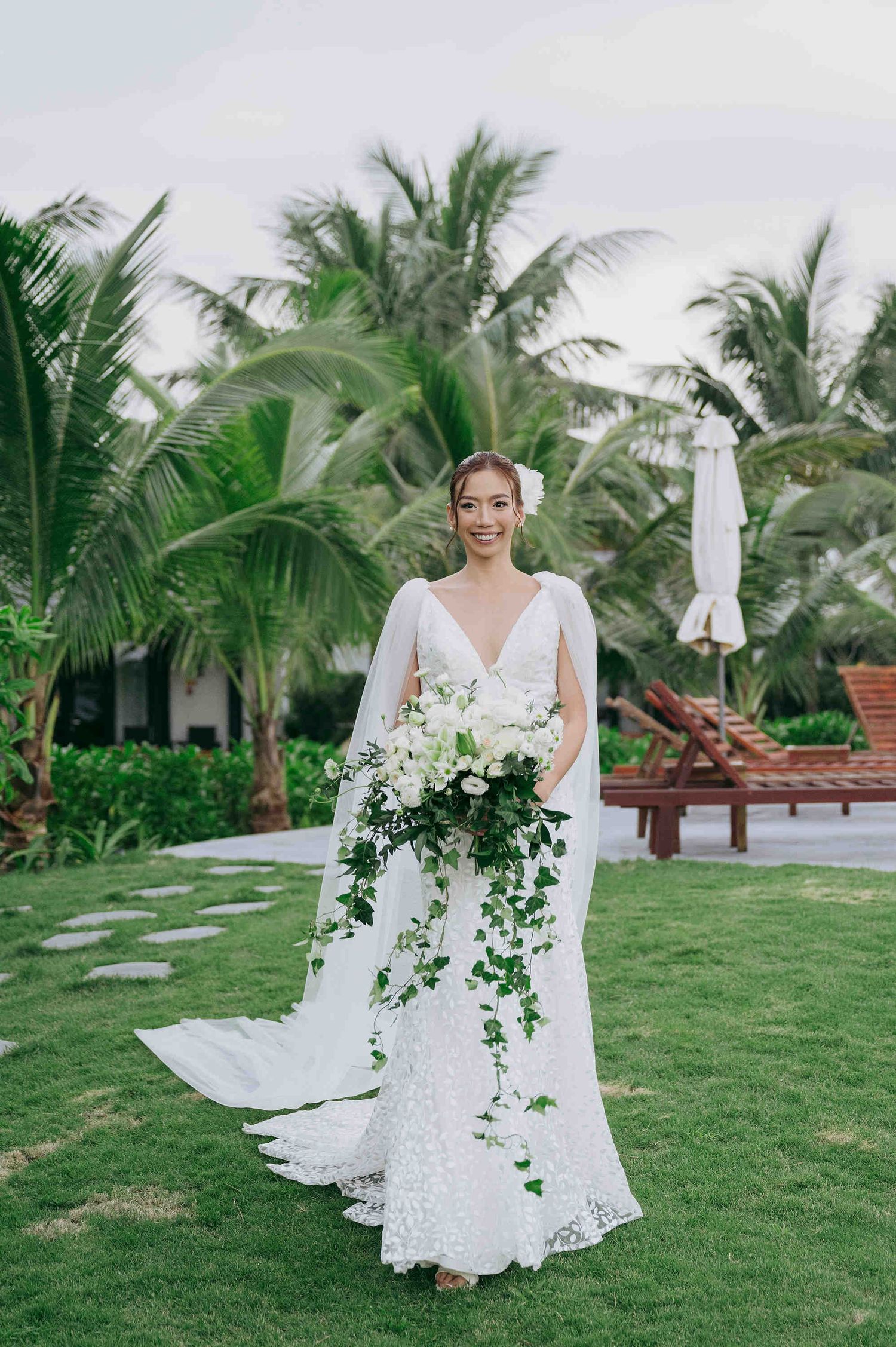 Bride in white lace gown with cape holds cascading green and white bouquet on tropical lawn with palm trees.