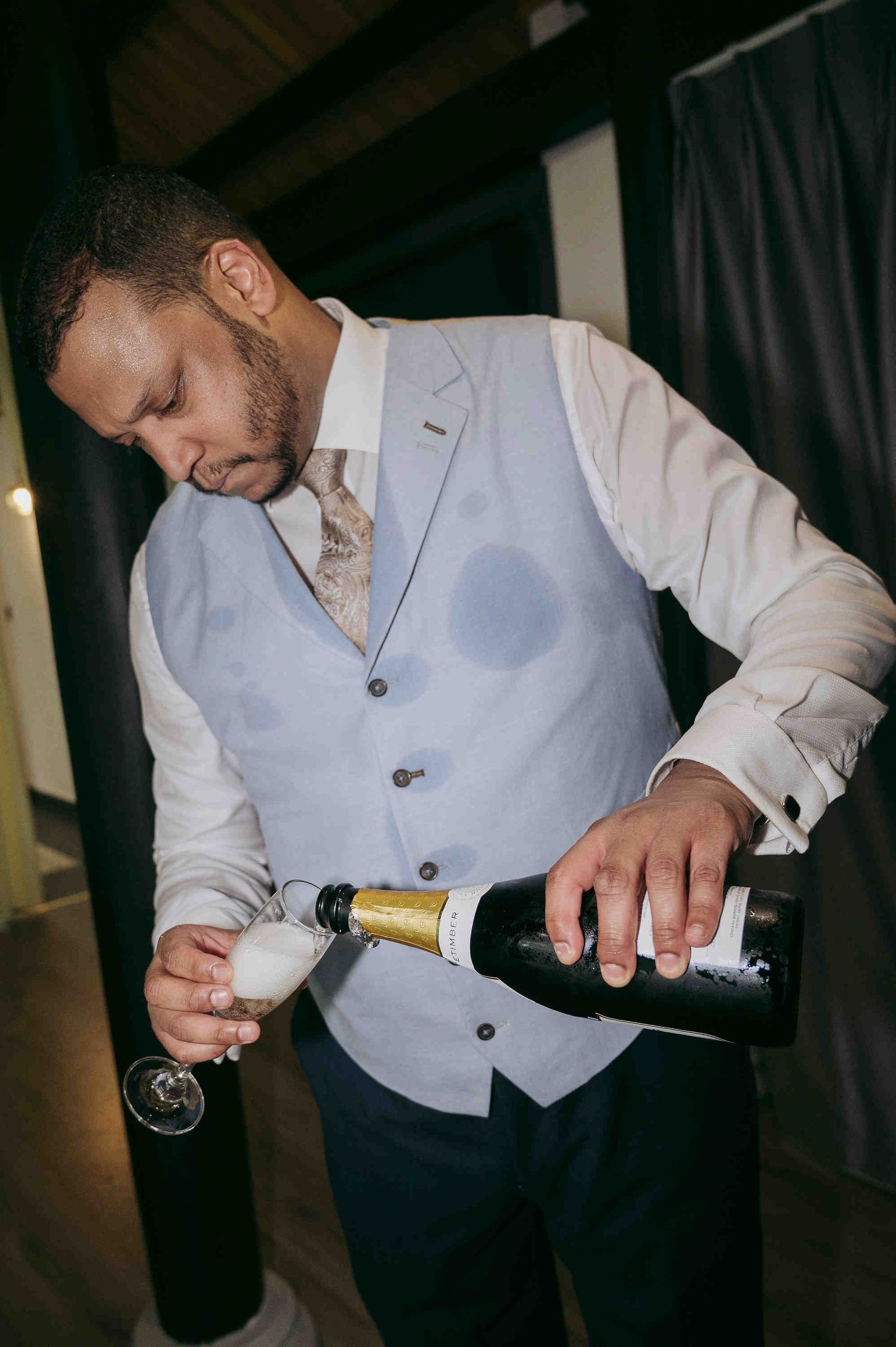 A well-dressed man in a blue vest pours champagne into a glass at an elegant event.