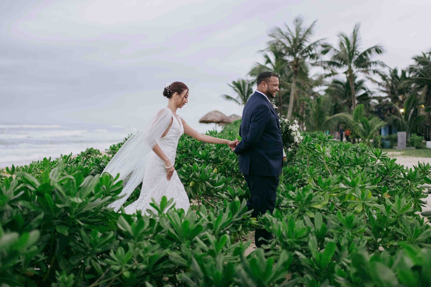 Bride and groom hold hands among lush tropical greenery with palm trees and ocean in background at beach wedding.