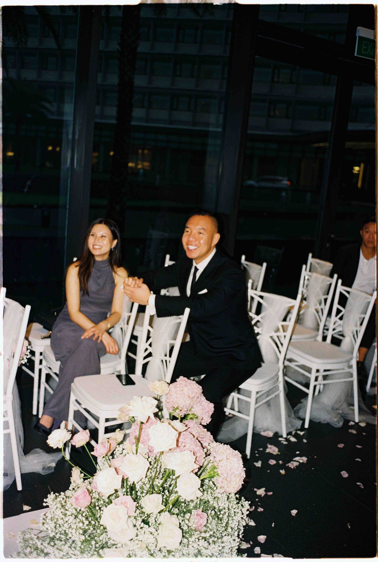 Da Nang wedding ceremony: couple smiling at wedding ceremony seated beside white chairs with pink floral arrangements, by AnhVuWedding.
