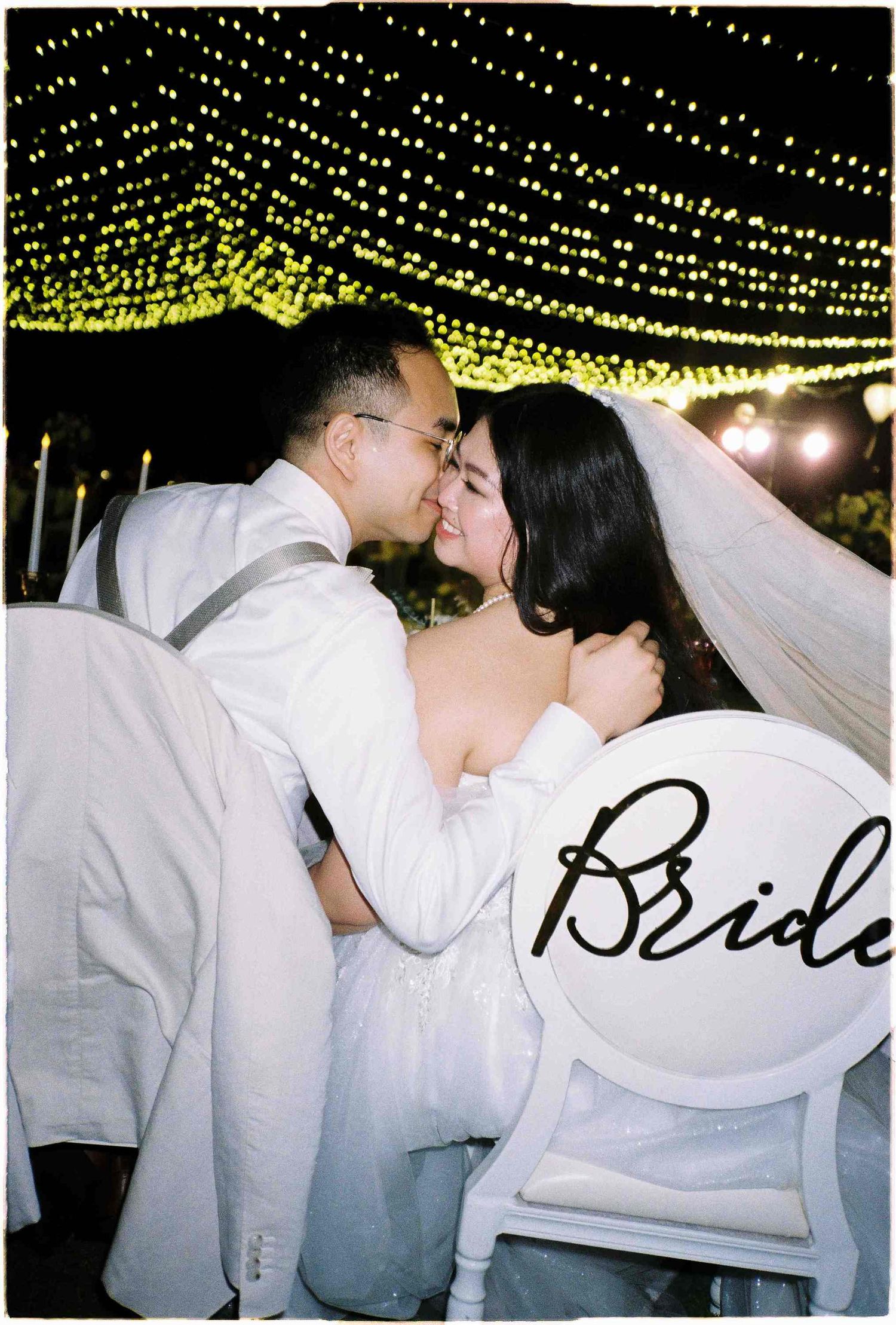 A bride and groom share a romantic kiss under twinkling fairy lights at their outdoor wedding reception.