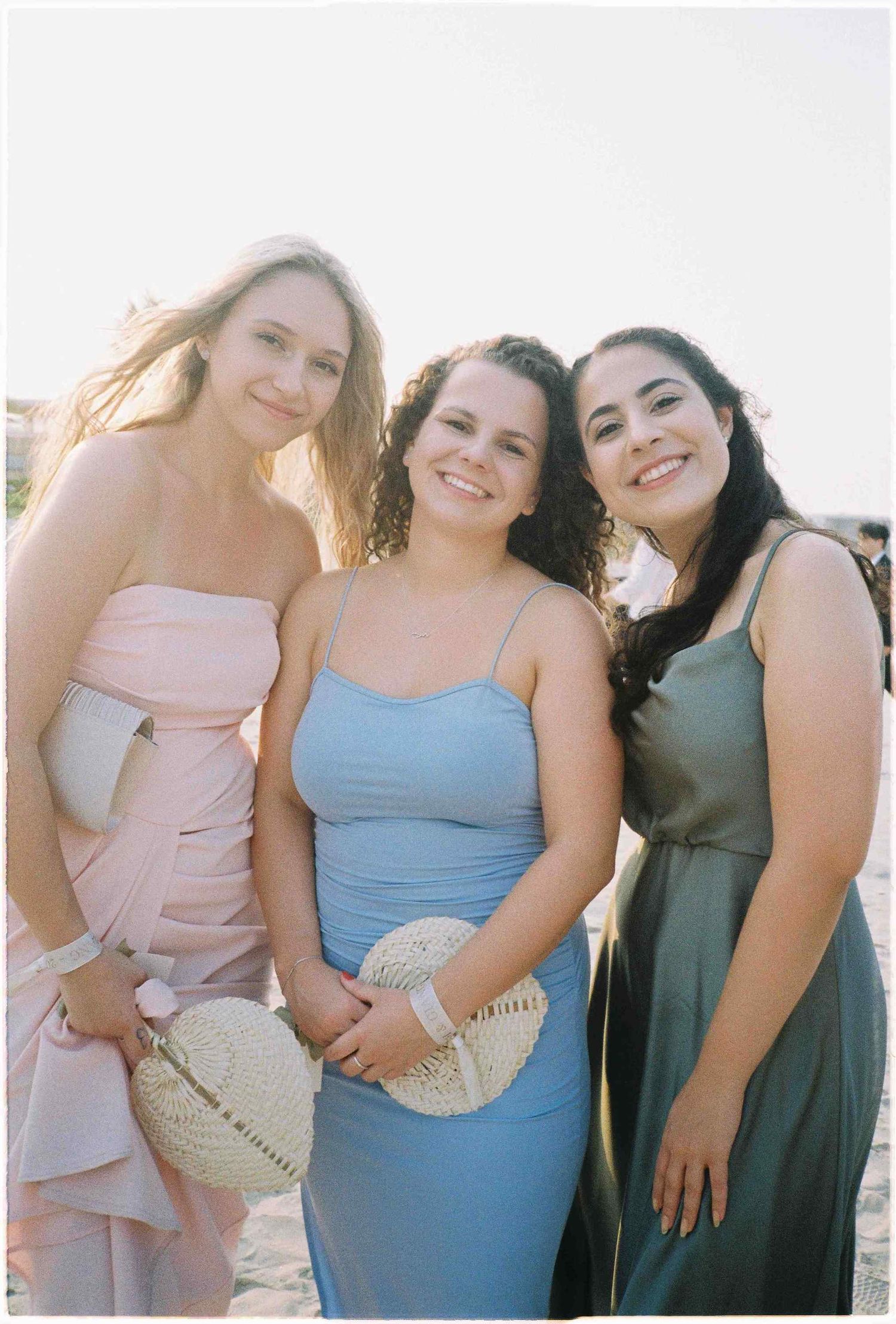 Three women in elegant dresses smile together at a beach event, wearing pink, blue, and gray gowns.