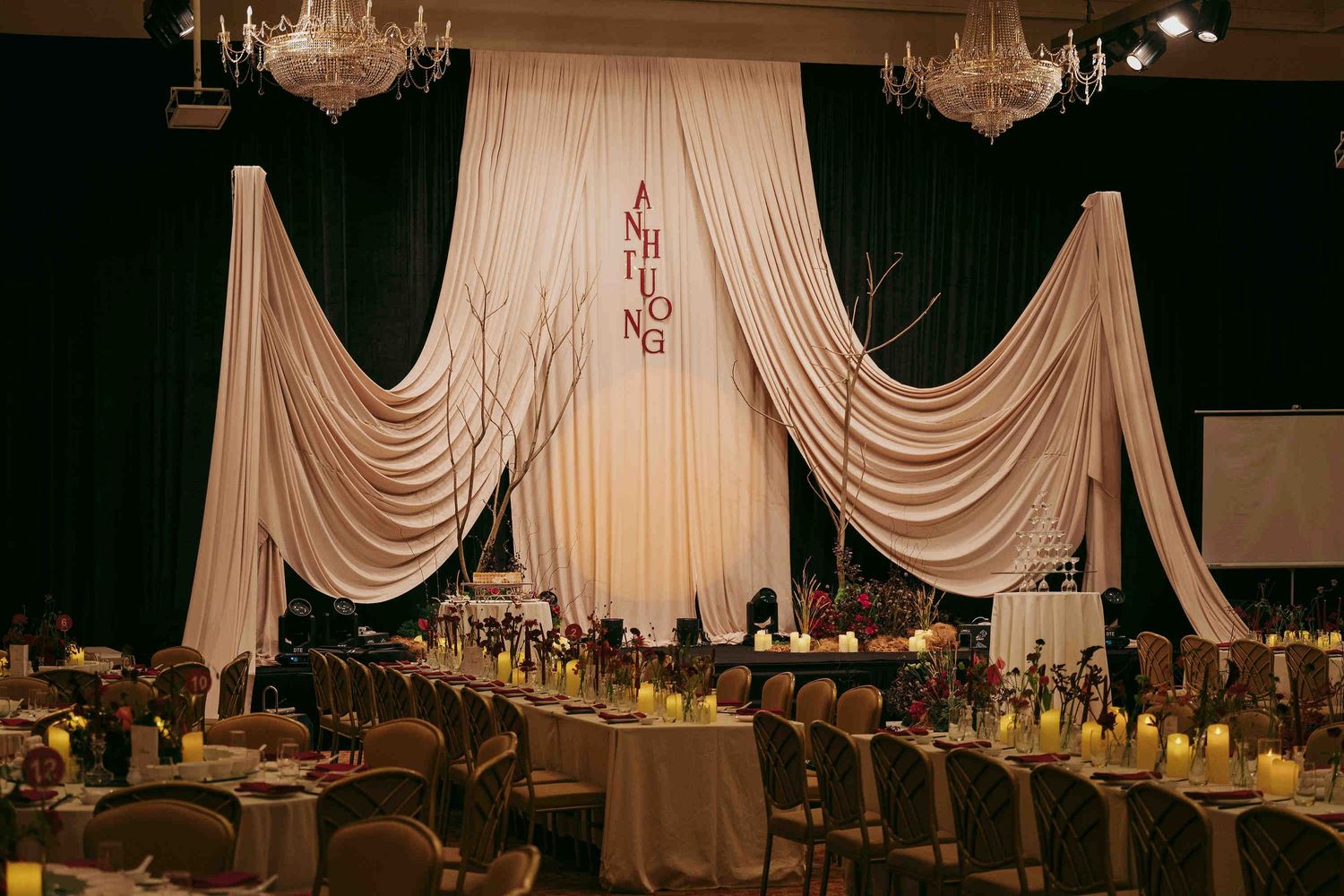 Elegant wedding reception hall with draped ivory fabric backdrop, monogram, chandelier lighting, and candlelit tables.