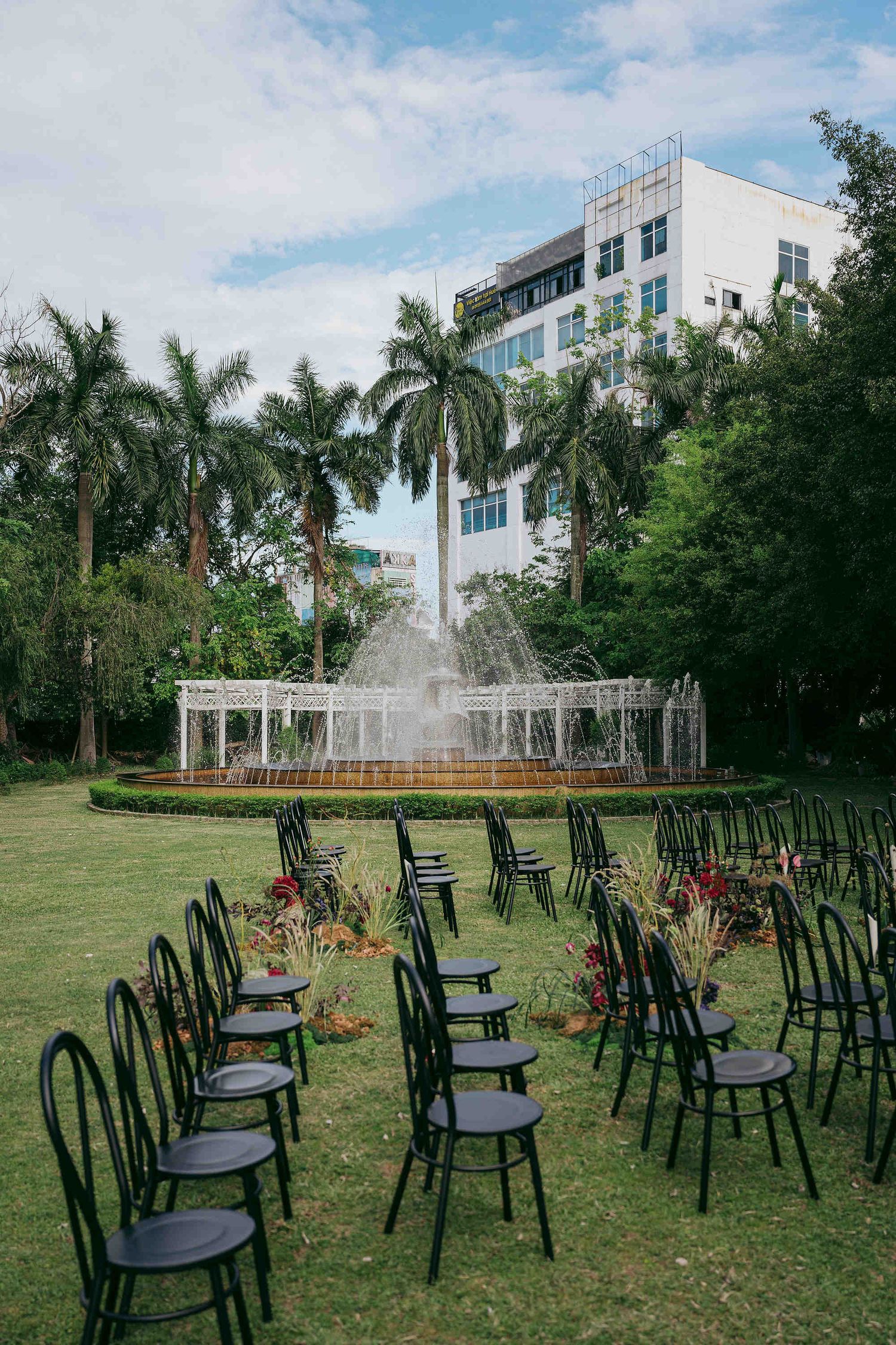 Black chairs arranged on green lawn facing ornate fountain with palm trees and white building backdrop for outdoor wedding ceremony.