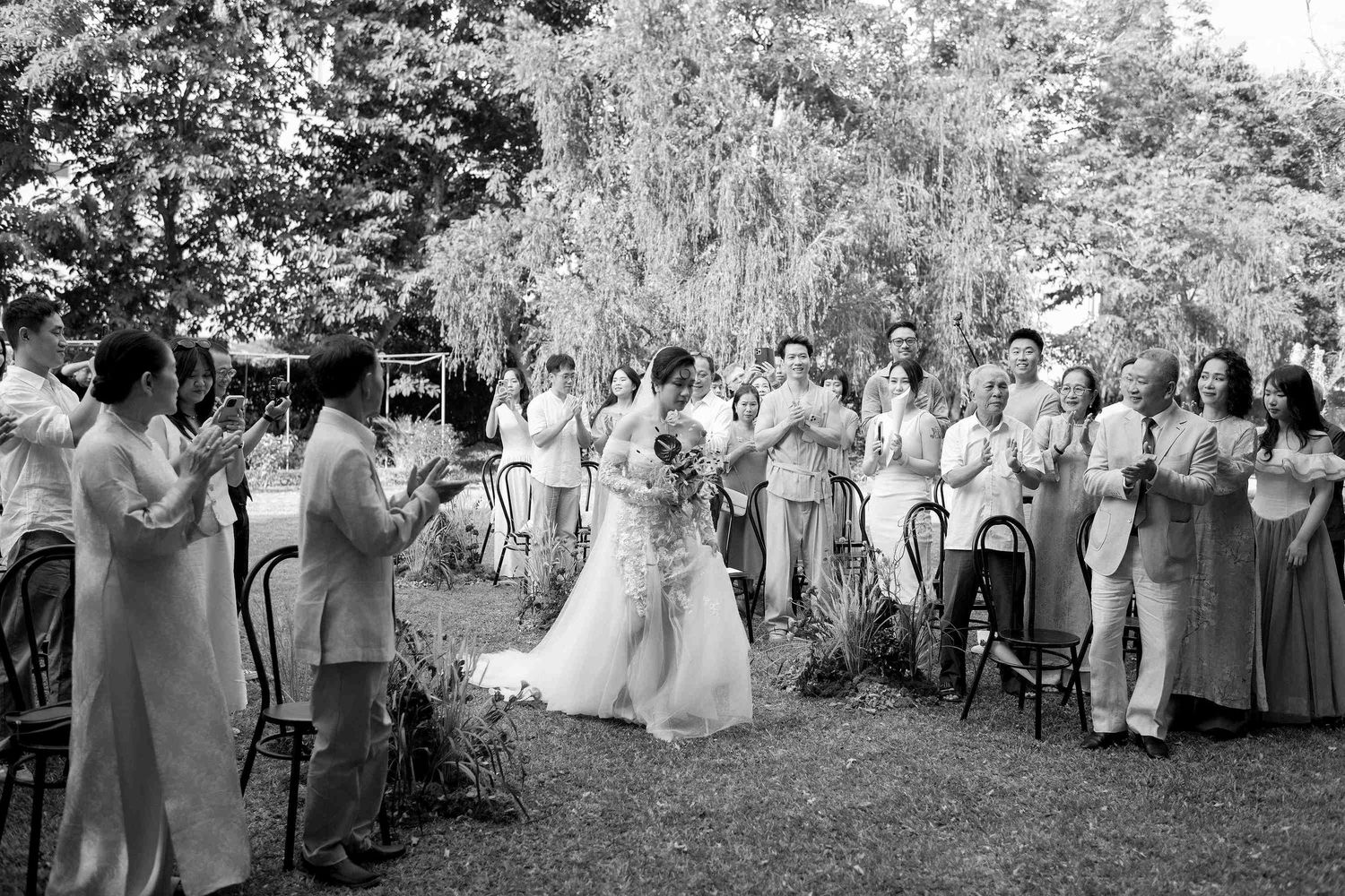 Black and white outdoor wedding ceremony with bride and groom surrounded by applauding guests on a lush garden lawn.