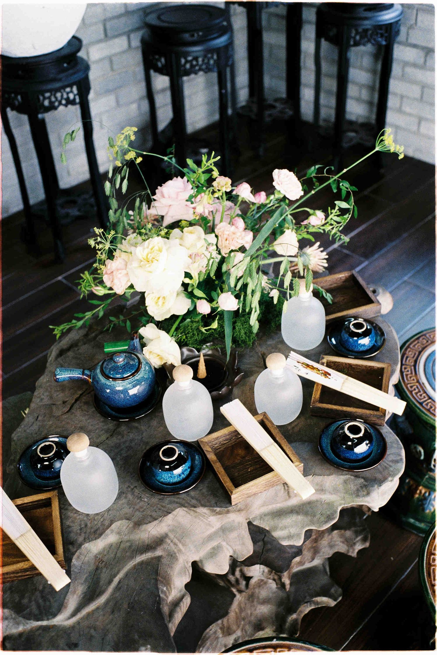 Elegant tea ceremony table with blue teapots, frosted glass vessels, wooden boxes, and pink floral centerpiece on stone surface.