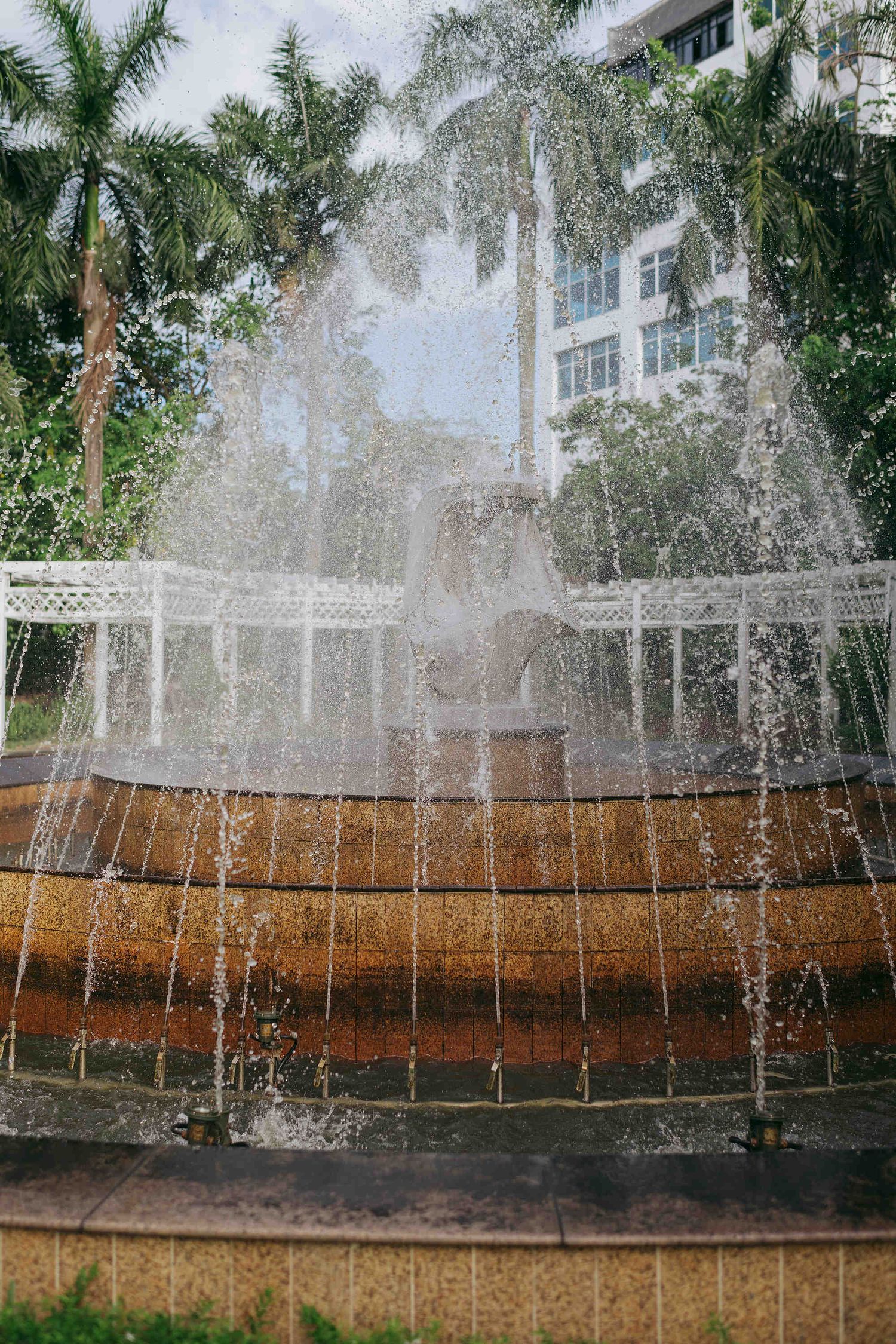 Ornate multi-tiered outdoor fountain with cascading water, surrounded by tropical palm trees and a white building.