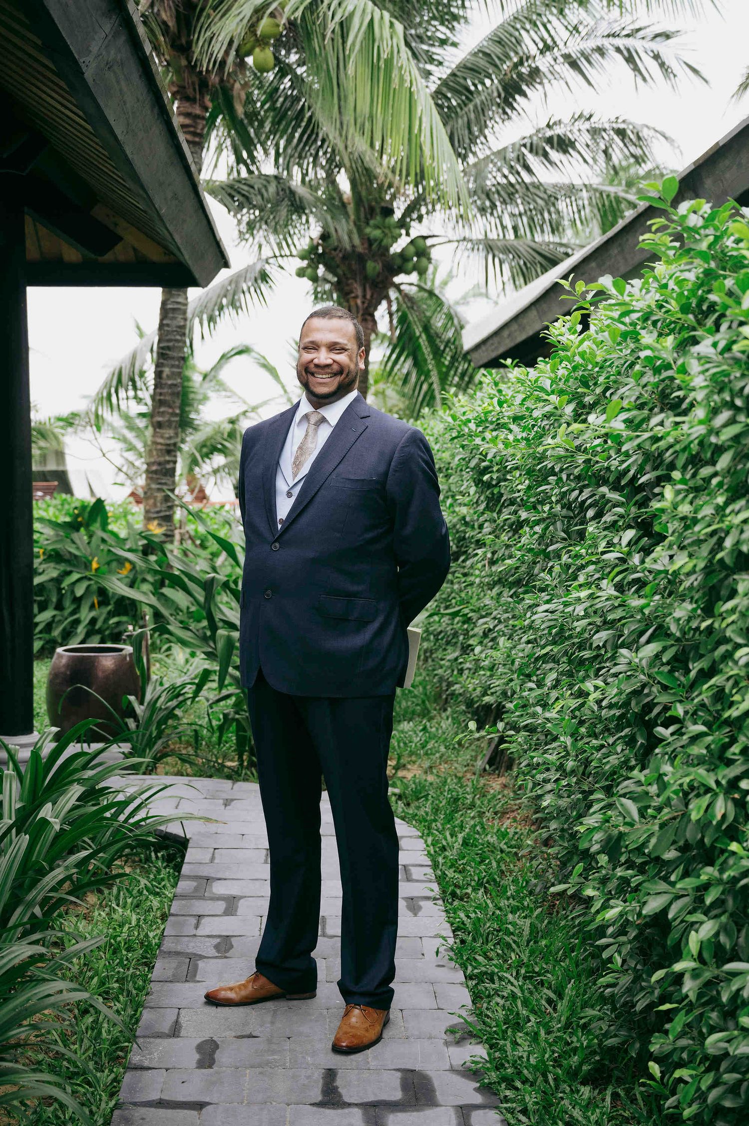 Smiling groom in navy suit and brown shoes standing on a garden path surrounded by tropical greenery.