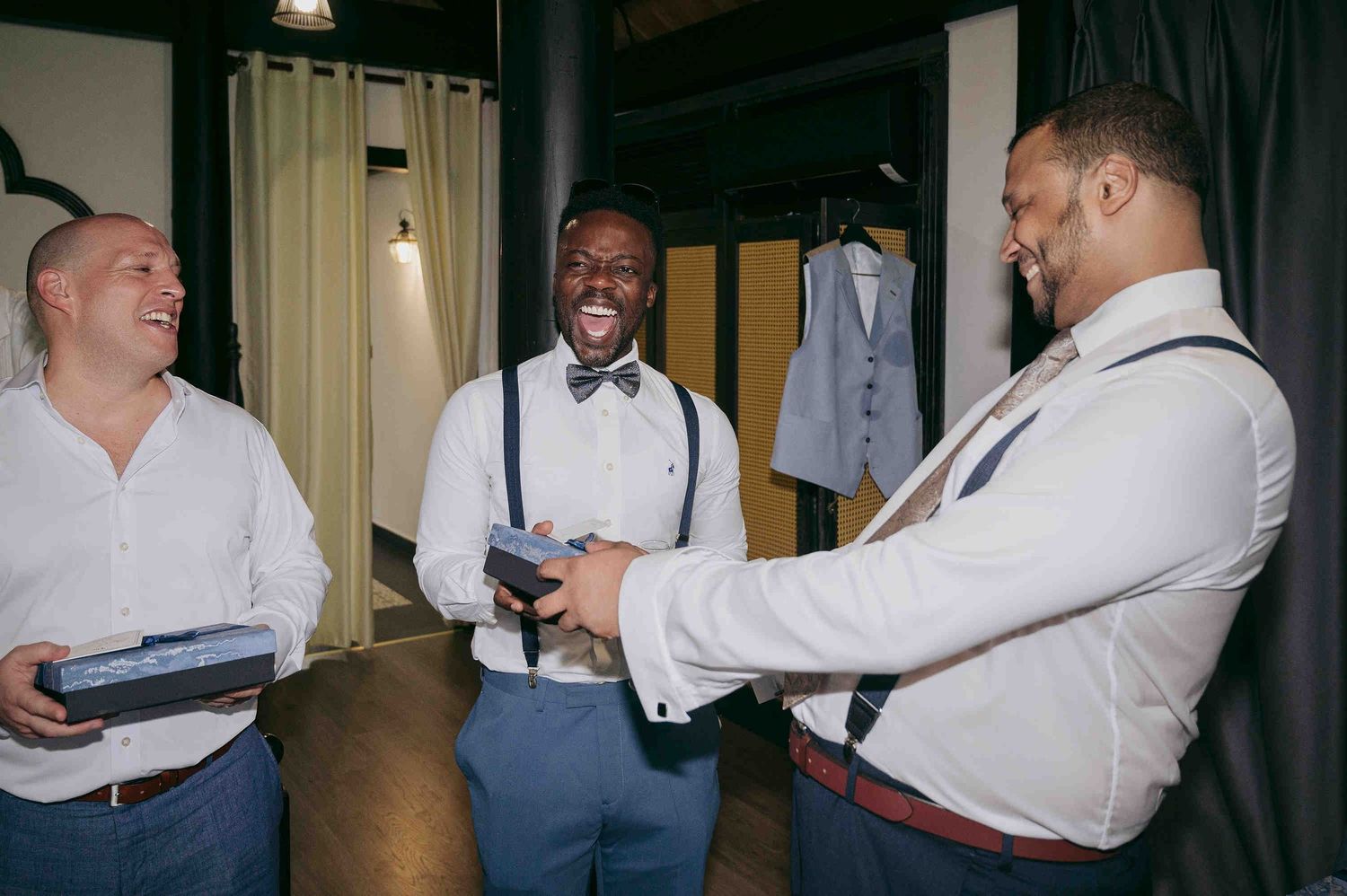 Three groomsmen in white shirts and suspenders laughing together while exchanging gifts before a wedding ceremony.