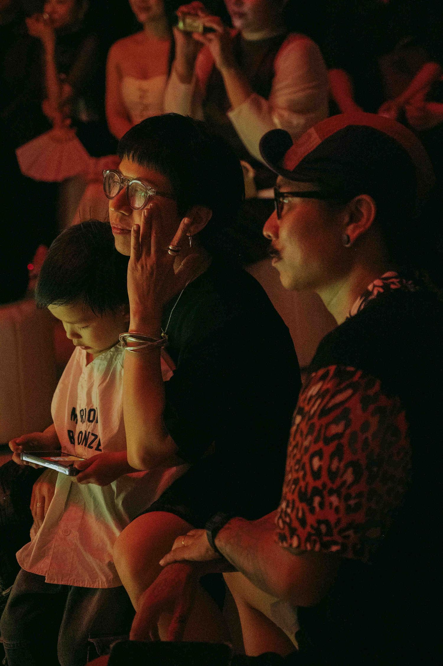 Audience members watch a performance under red lighting, with one person wearing glasses and a leopard print shirt.