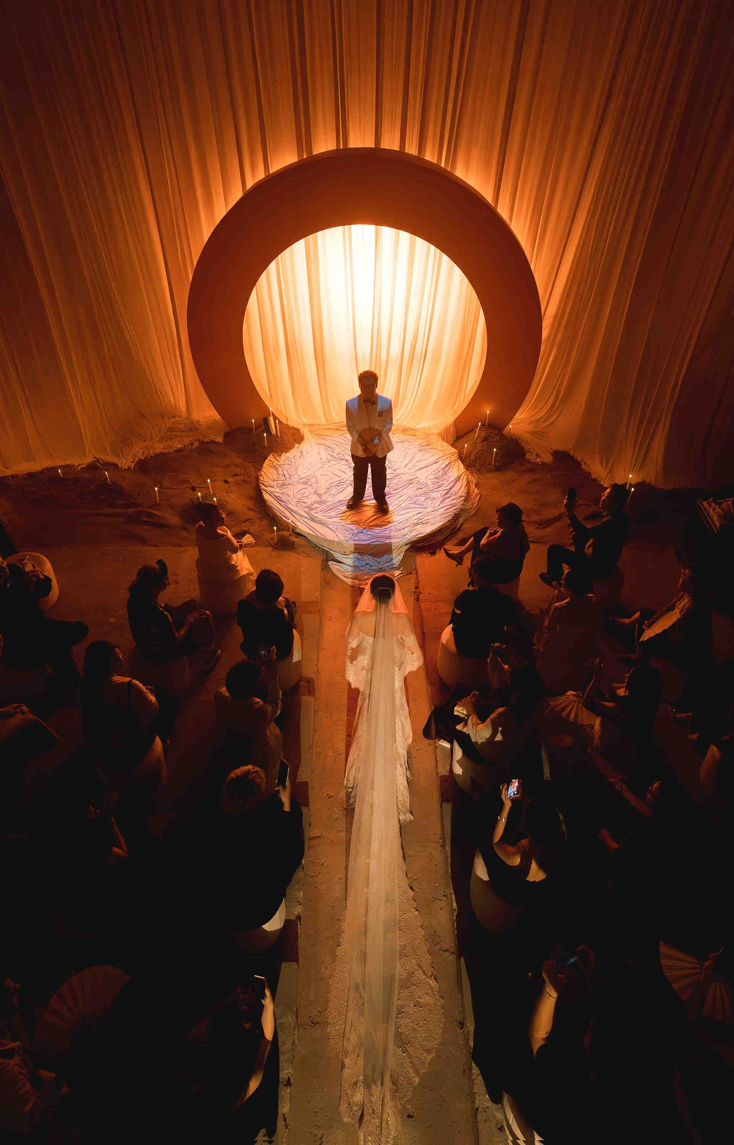 Bride walking down aisle toward glowing circular arch with officiant, surrounded by guests in warm amber-lit wedding venue.