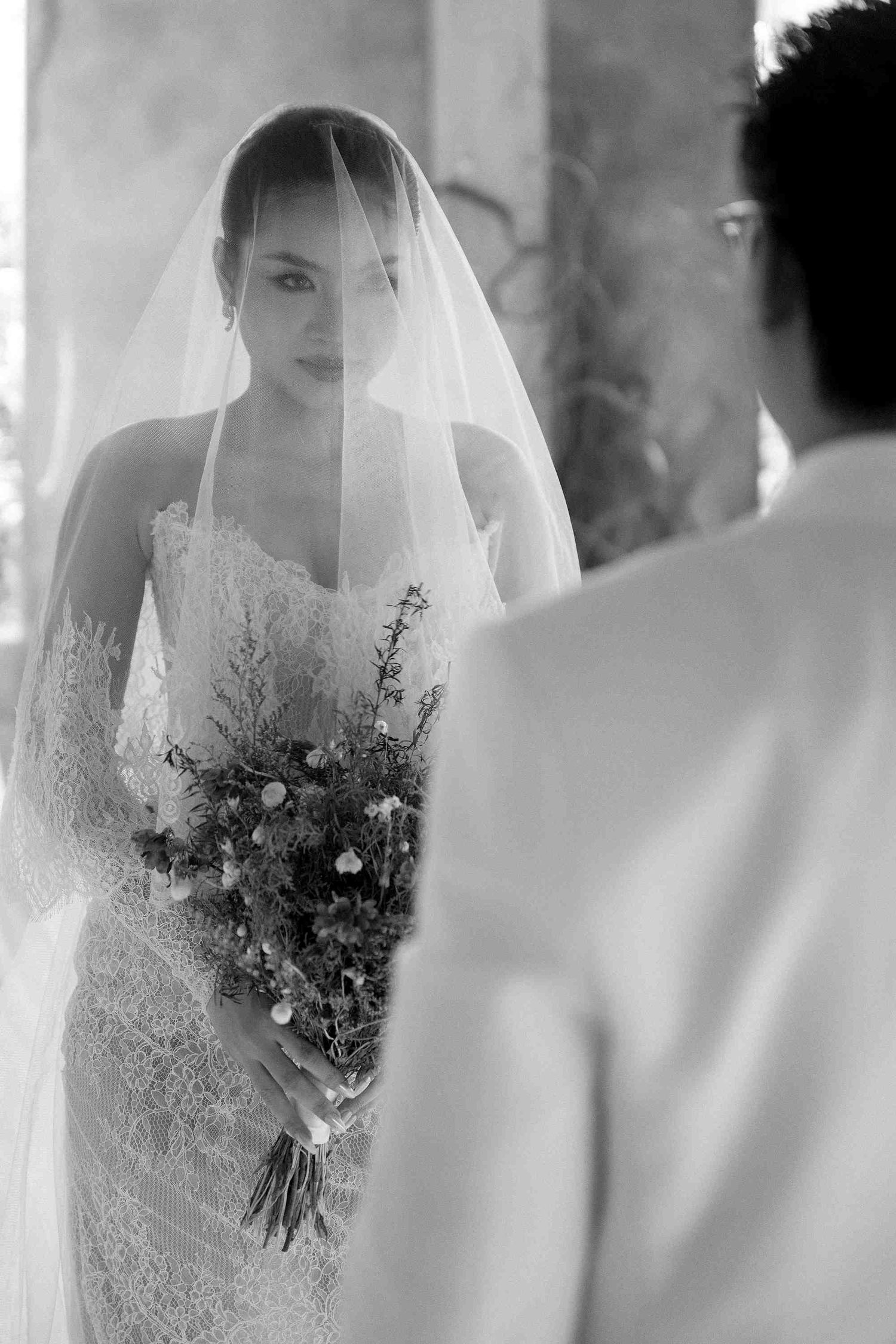 Black and white photo of a bride in lace gown and veil holding a bouquet, facing her groom at a wedding ceremony.