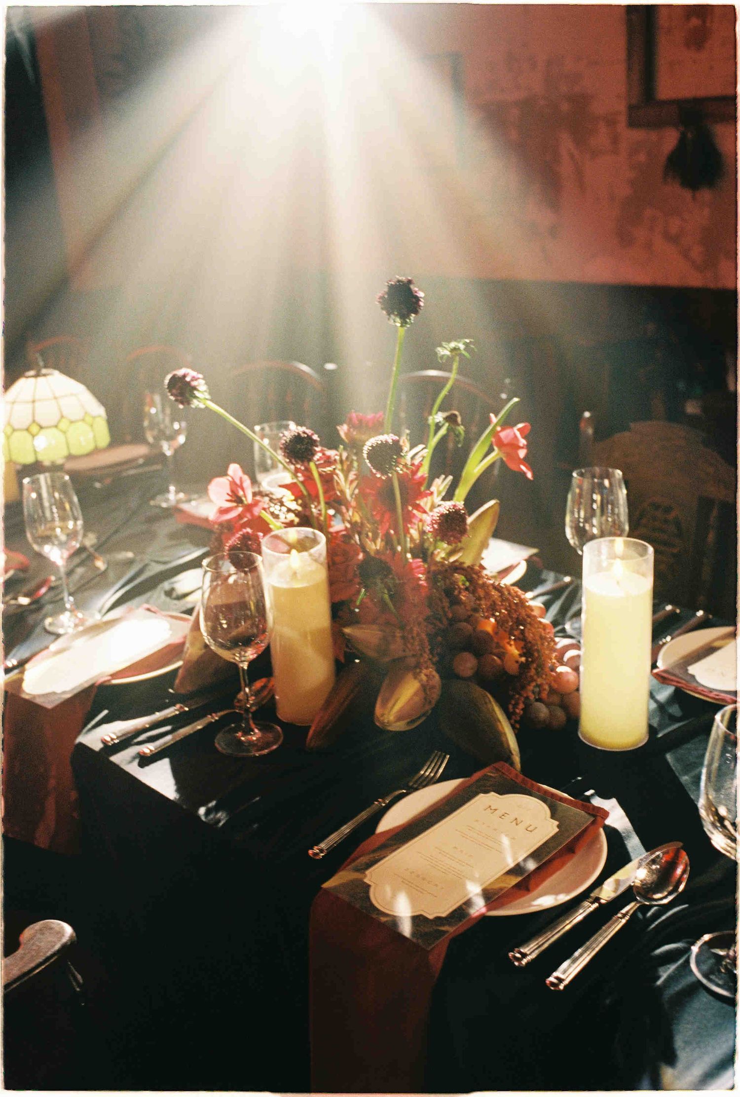 Close-up of a formal dinner table with floral centerpiece, candles, wine glasses, and place settings in warm light.