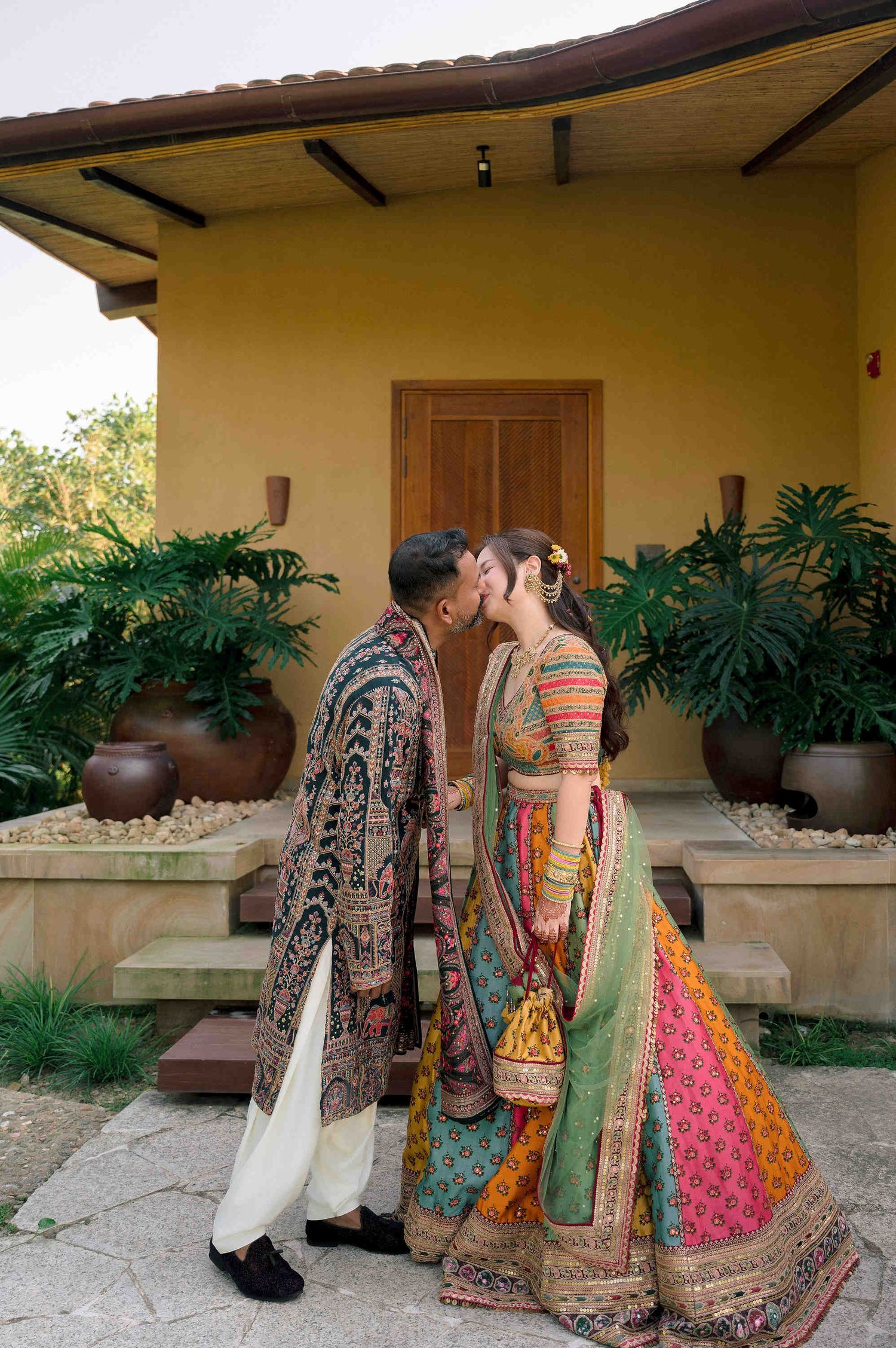 Couple in vibrant traditional Indian wedding attire sharing a kiss outside a yellow villa surrounded by lush greenery.