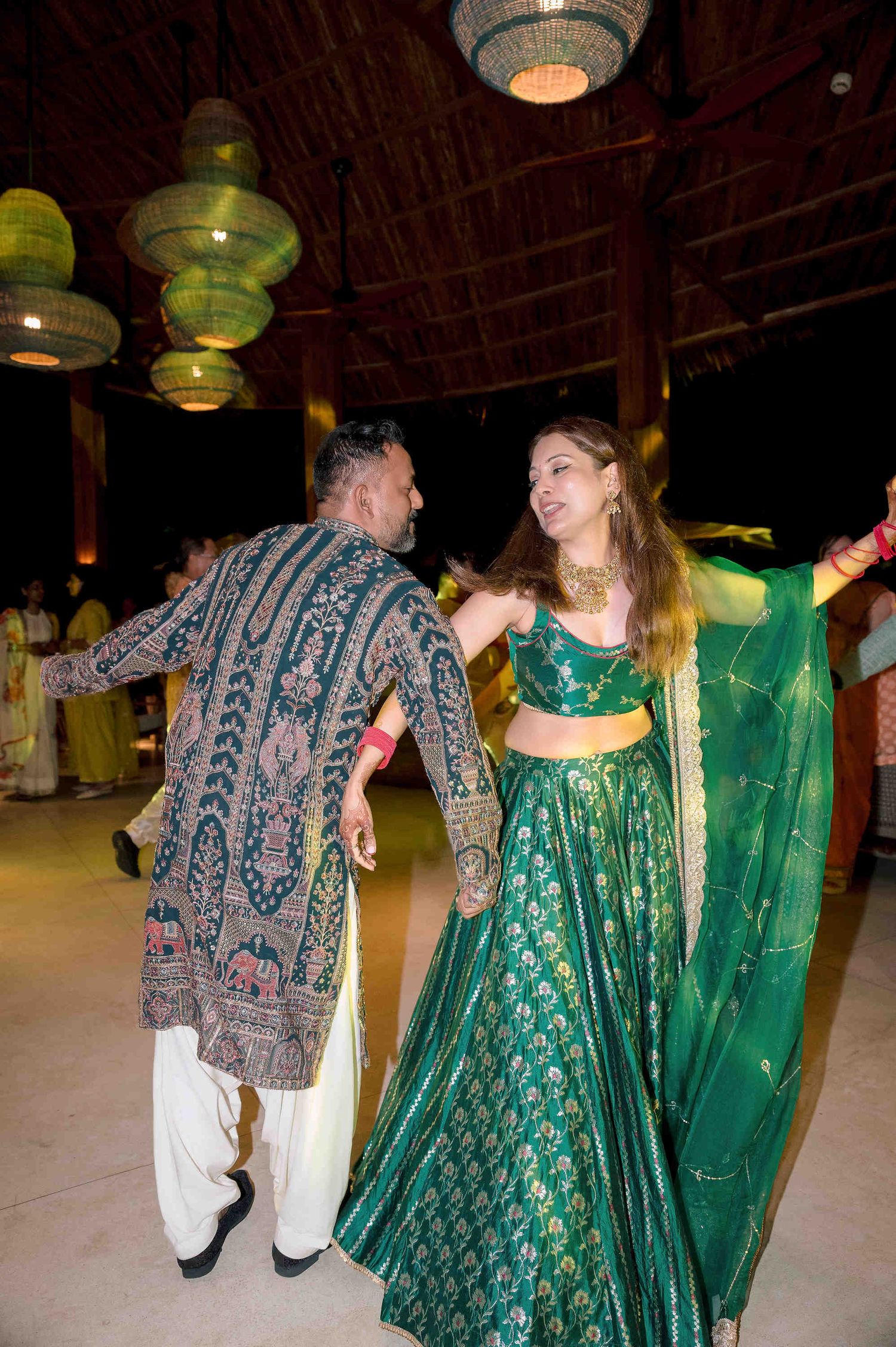 Couple dancing at Indian wedding celebration, woman in green lehenga, man in embroidered sherwani, elegant venue with hanging lanterns.