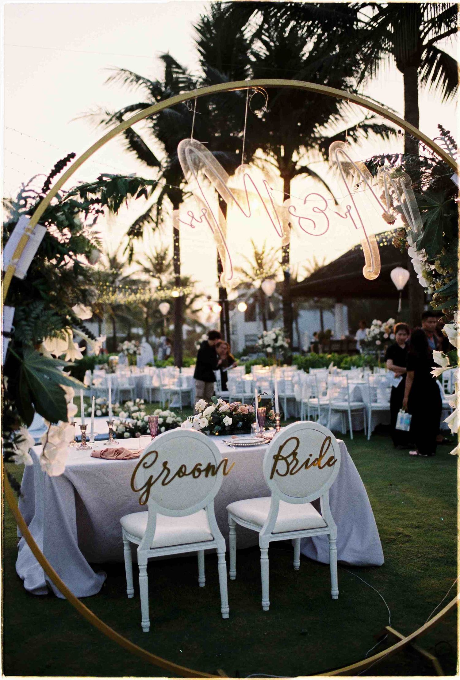 Elegant outdoor wedding reception with bride and groom chairs under a golden hoop arch with neon sign at sunset.