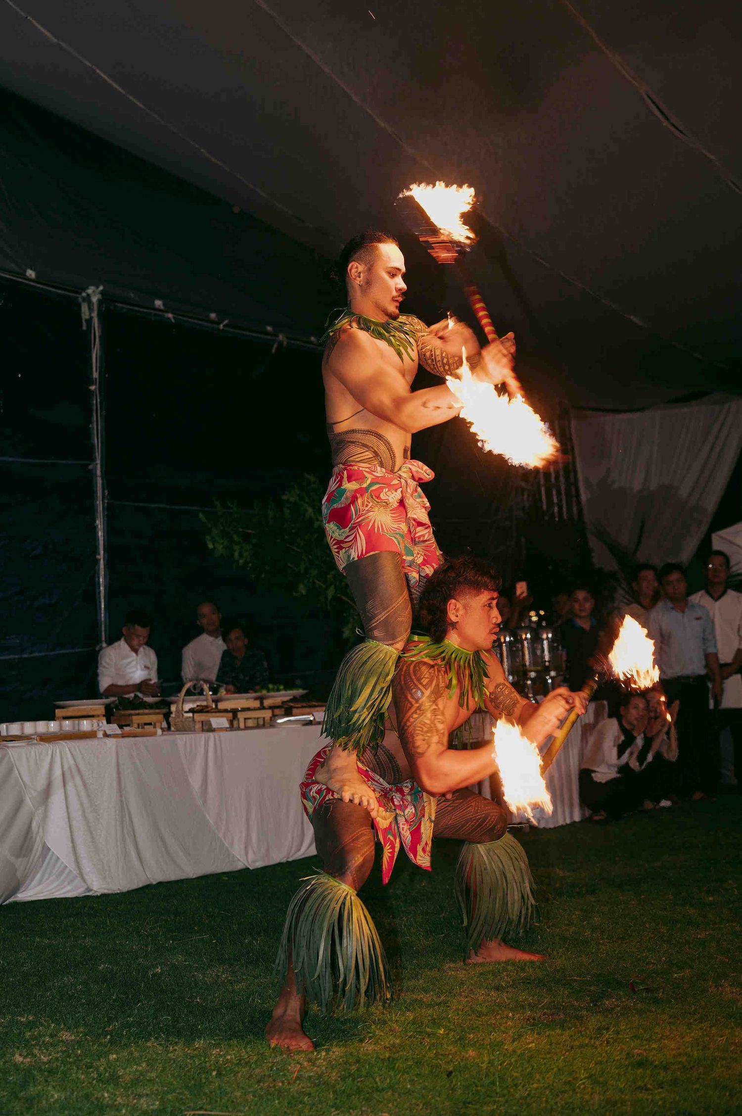 Two Polynesian fire dancers perform acrobatic routine at outdoor luau event, one standing on shoulders holding flaming torches.