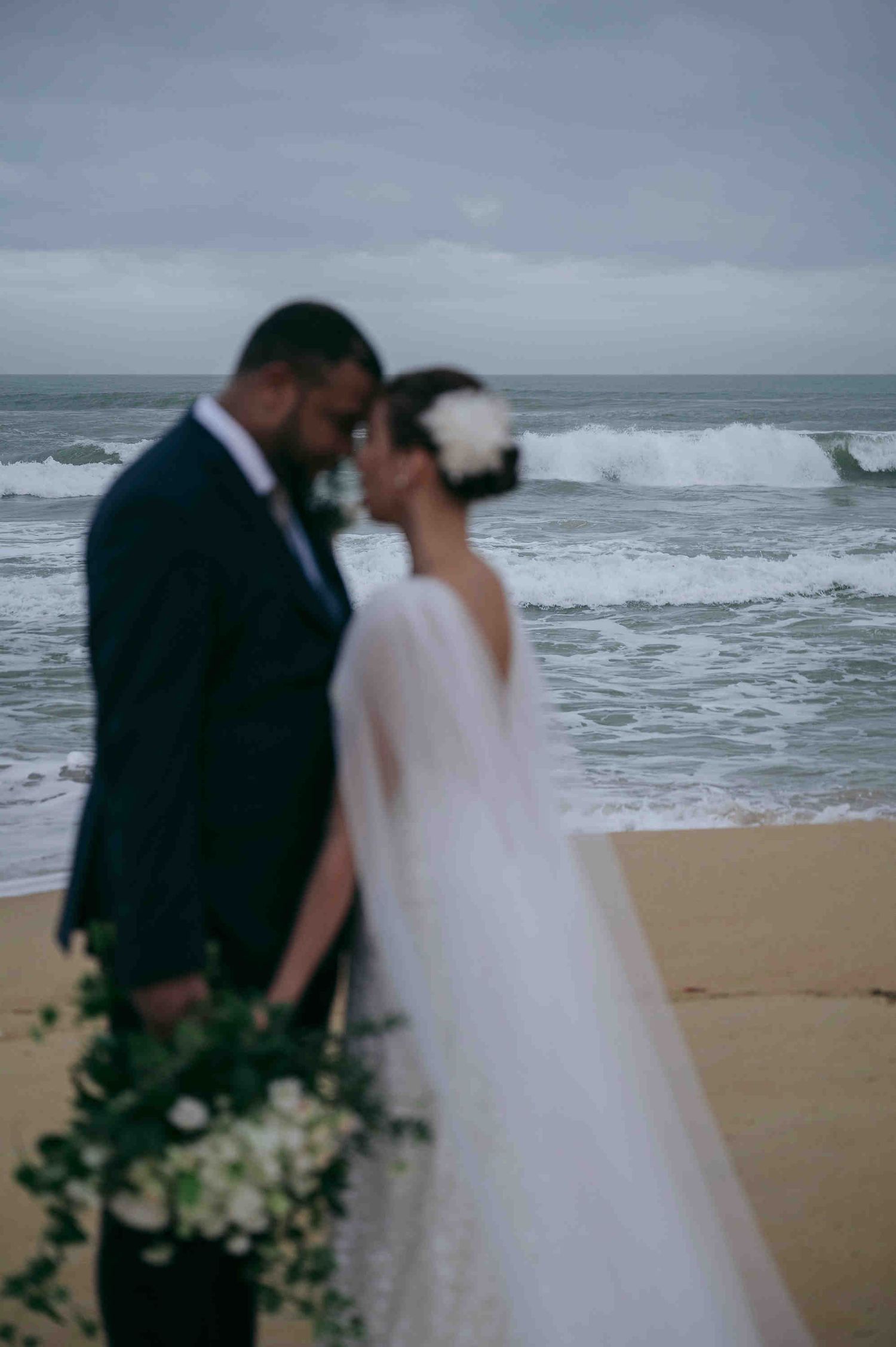 Bride and groom share an intimate moment on a sandy beach with dramatic ocean waves in the background on their wedding day.