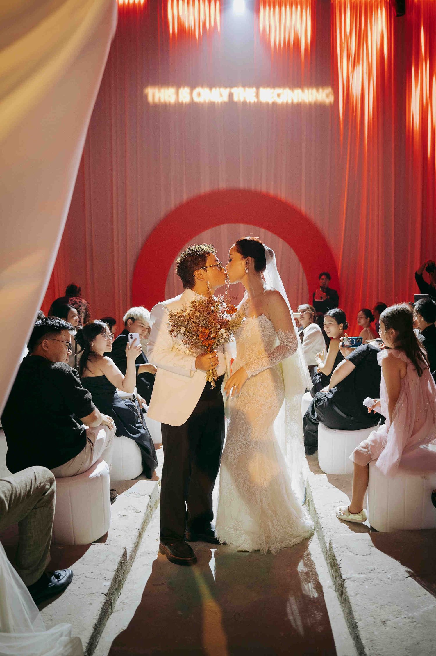 Bride and groom share first kiss at elegant wedding ceremony with red arch backdrop and glowing neon sign.