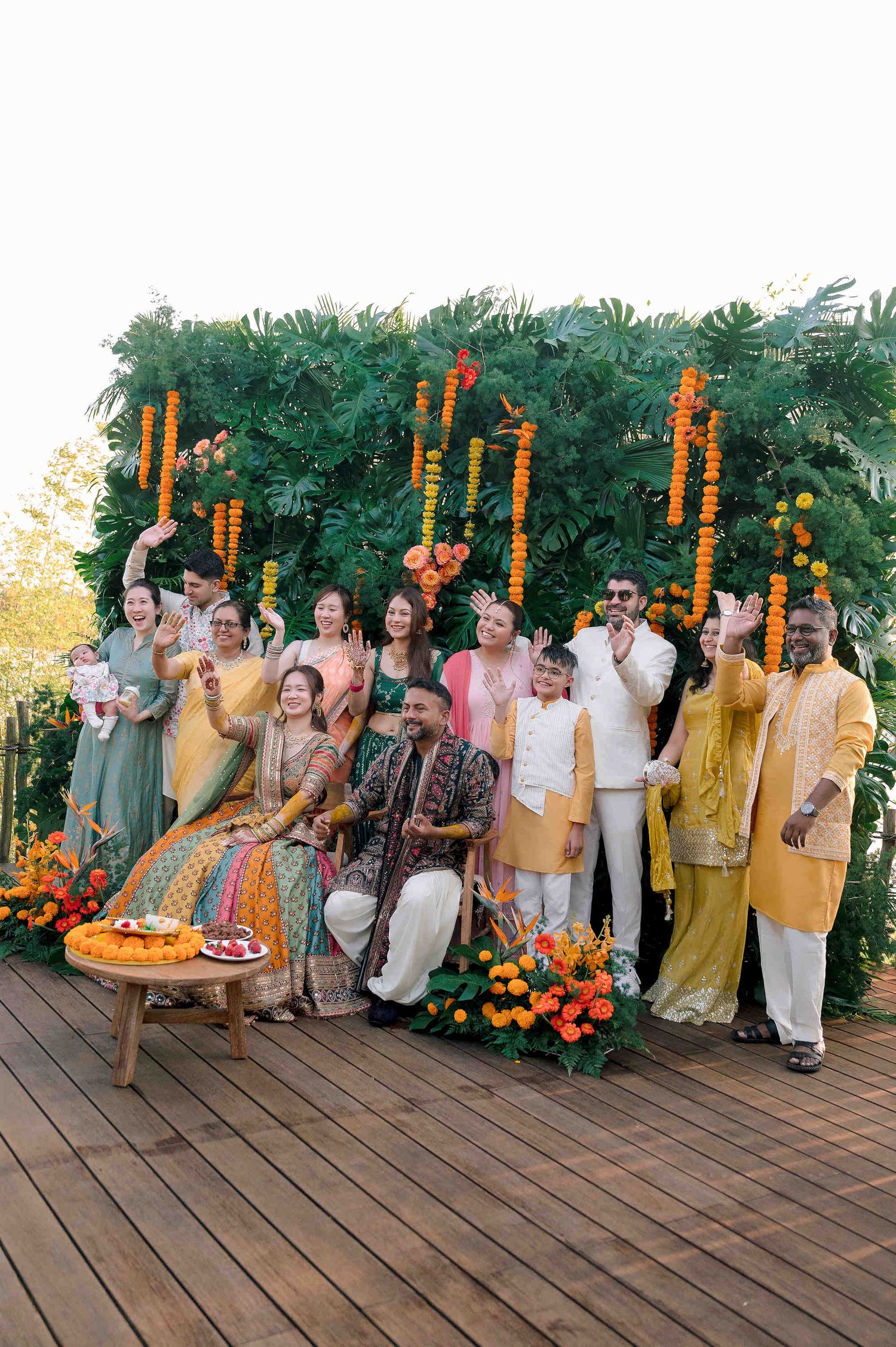 Indian wedding group photo with bride and groom in colorful attire, surrounded by family on a deck with marigold garland backdrop.